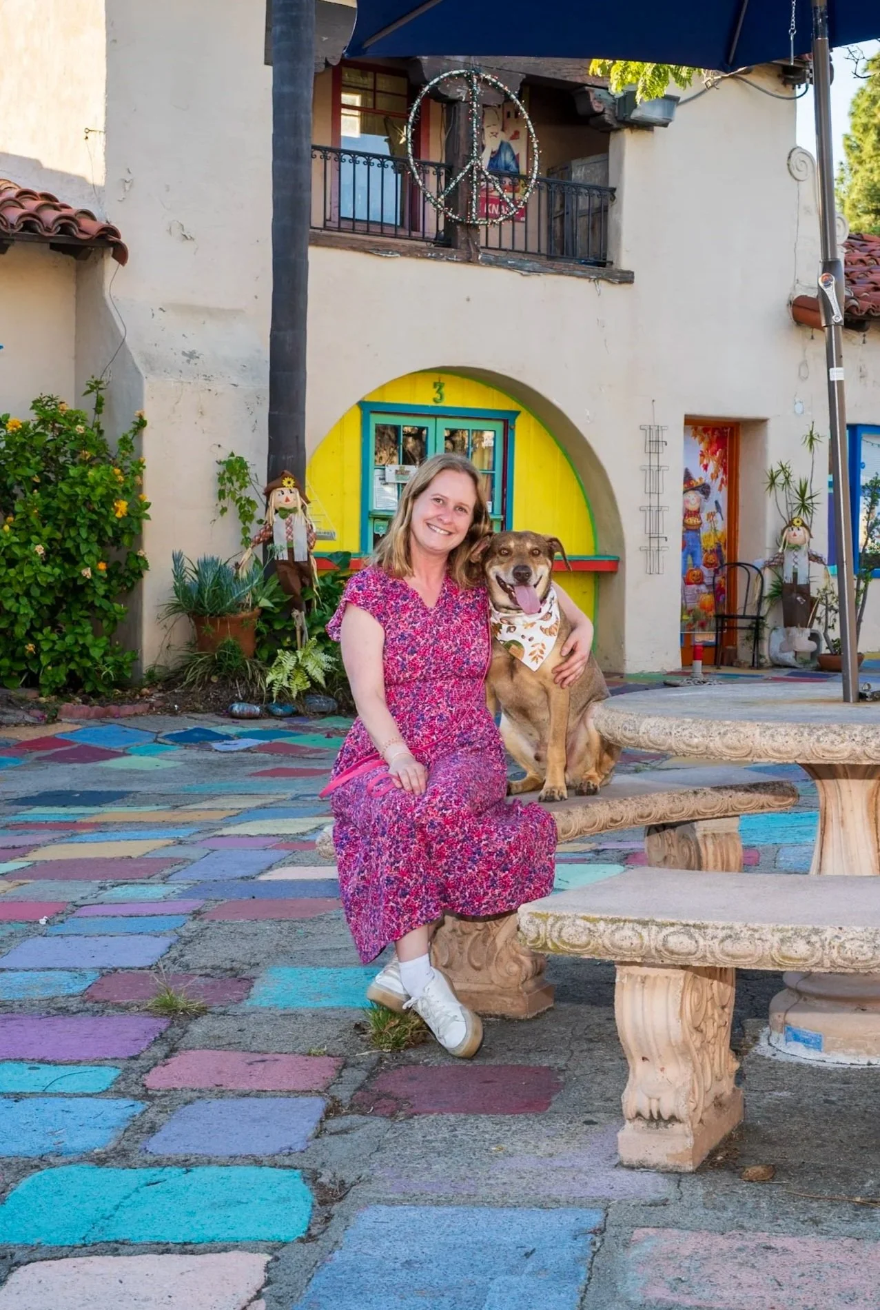 A woman in a pink floral dress sitting on a stone bench with a large brown dog wearing a bandana, in front of a colorful mural and decorated building.