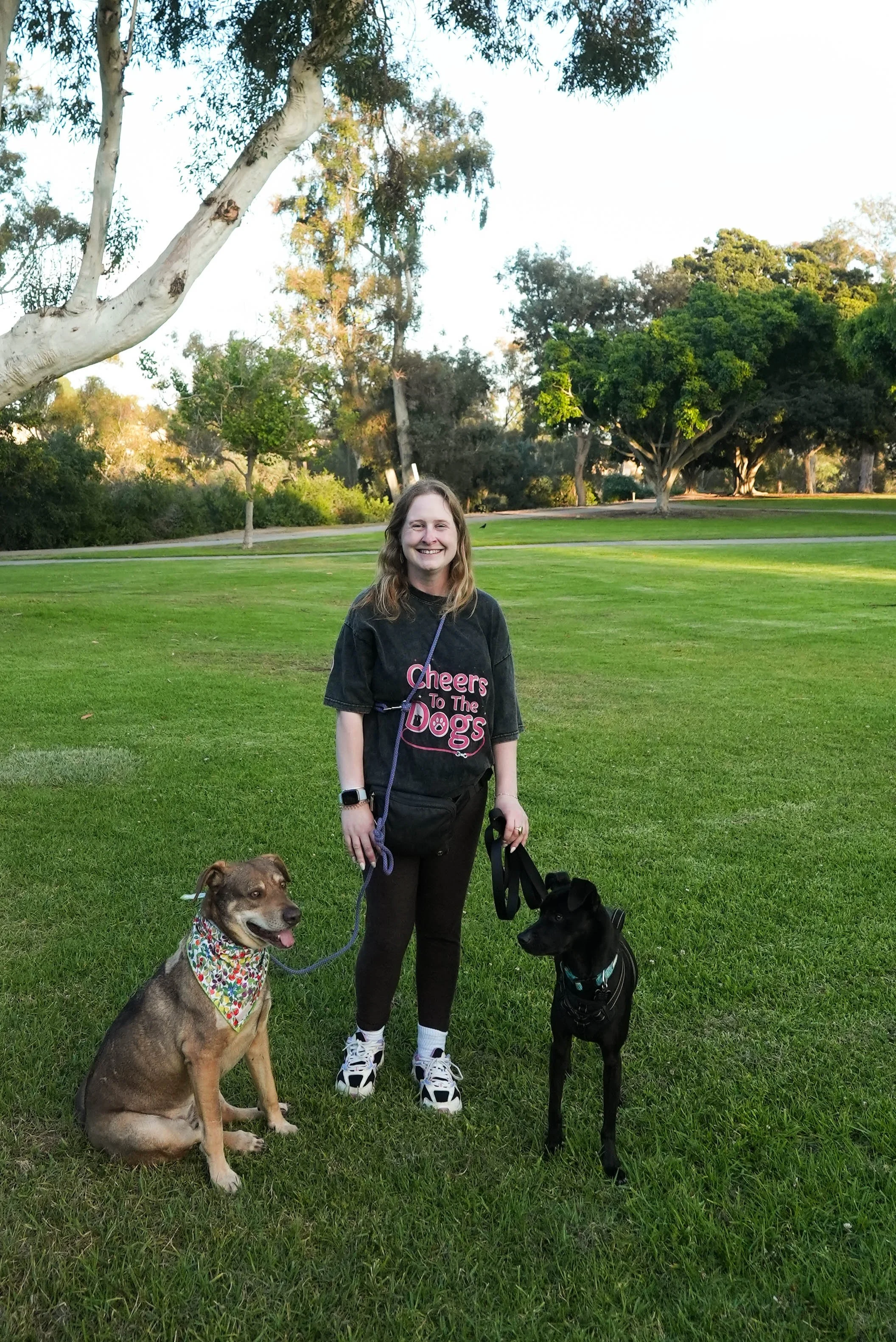 A woman with long hair, smiling, wearing a black t-shirt with pink text that says "Cheers to the Dogs," and black pants, standing on a grassy park with lush trees in the background. She is holding two leash cords leading to two dogs: a sitting brown and black dog with a colorful bandana around its neck, and a standing black dog with a harness, both on leash.