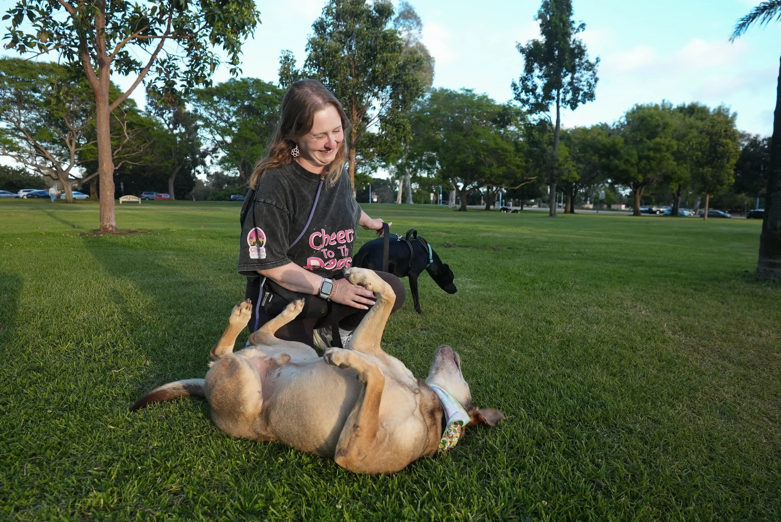 A woman wearing a black shirt with pink text and earrings, smiling while petting a dog. The dog is lying on its back on the grass with its legs up, wearing a bandana. The background shows a park with trees and parked cars.