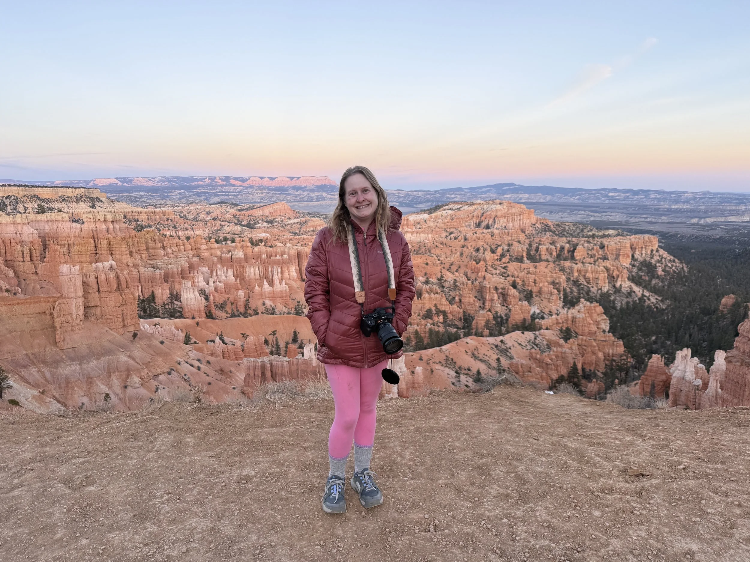 Woman standing on the edge of a canyon with red rock formations, wearing pink leggings and a red jacket, with a camera hanging around her neck, during sunset.