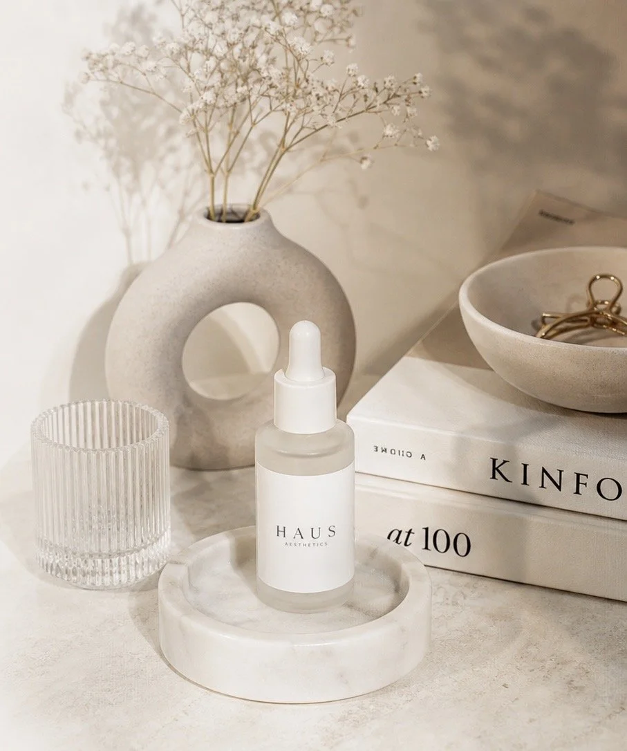 A minimalist arrangement of decorative items on a light-colored surface, including a white dropper bottle labeled "HAUS AESTHETICS," a clear ribbed glass, a beige vase with dried white flowers, a stack of art books titled "KIN F A T 100," a beige ceramic dish with gold jewelry, and a round white marble tray.