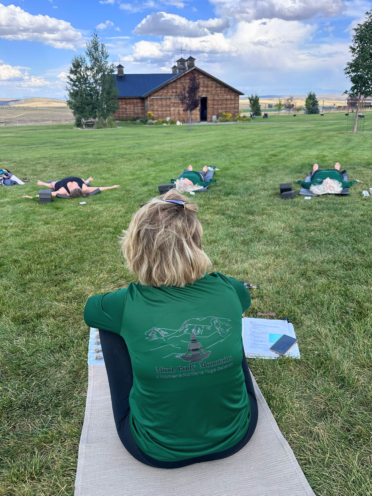 Daneesha overlooking retreat participants enjoying an outdoor yoga class in a grassy field, with a rustic barn and landscape in the background, under a partly cloudy sky.