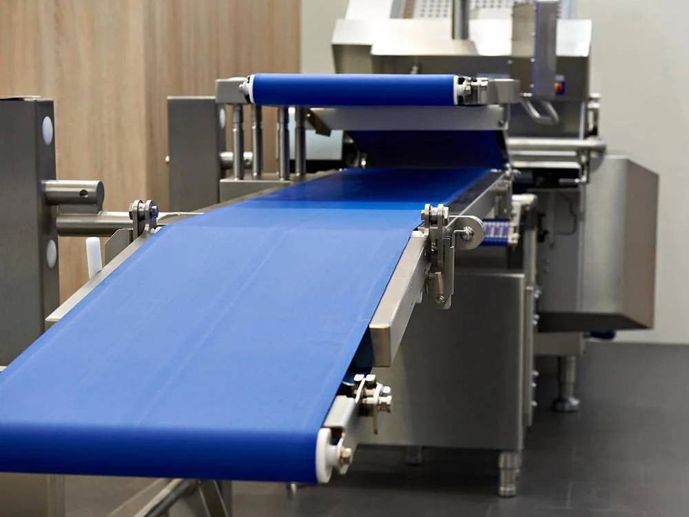 Máquina industrial con banda transportadora azul en un entorno de fábrica o laboratorio.