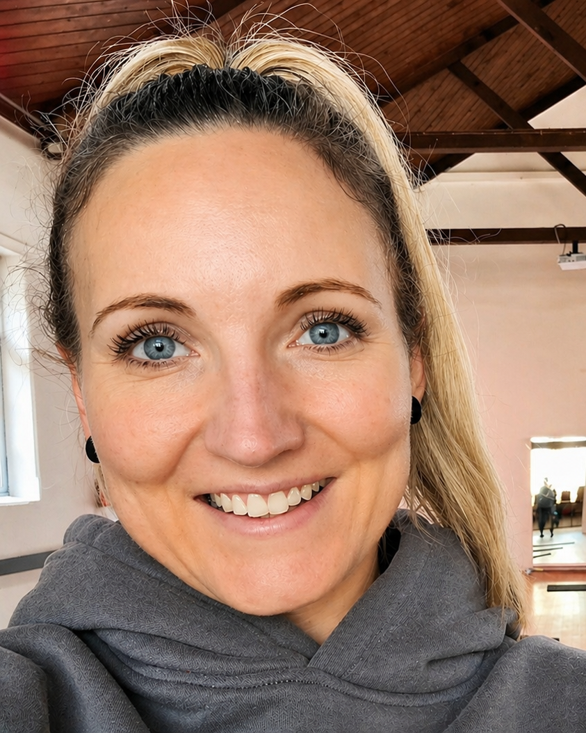 Close-up of a smiling female Pilates Instructor with blonde hair pulled back in a ponytail, blue eyes, and wearing a gray hoodie, inside a church hall with a wooden ceiling and a window in the background.