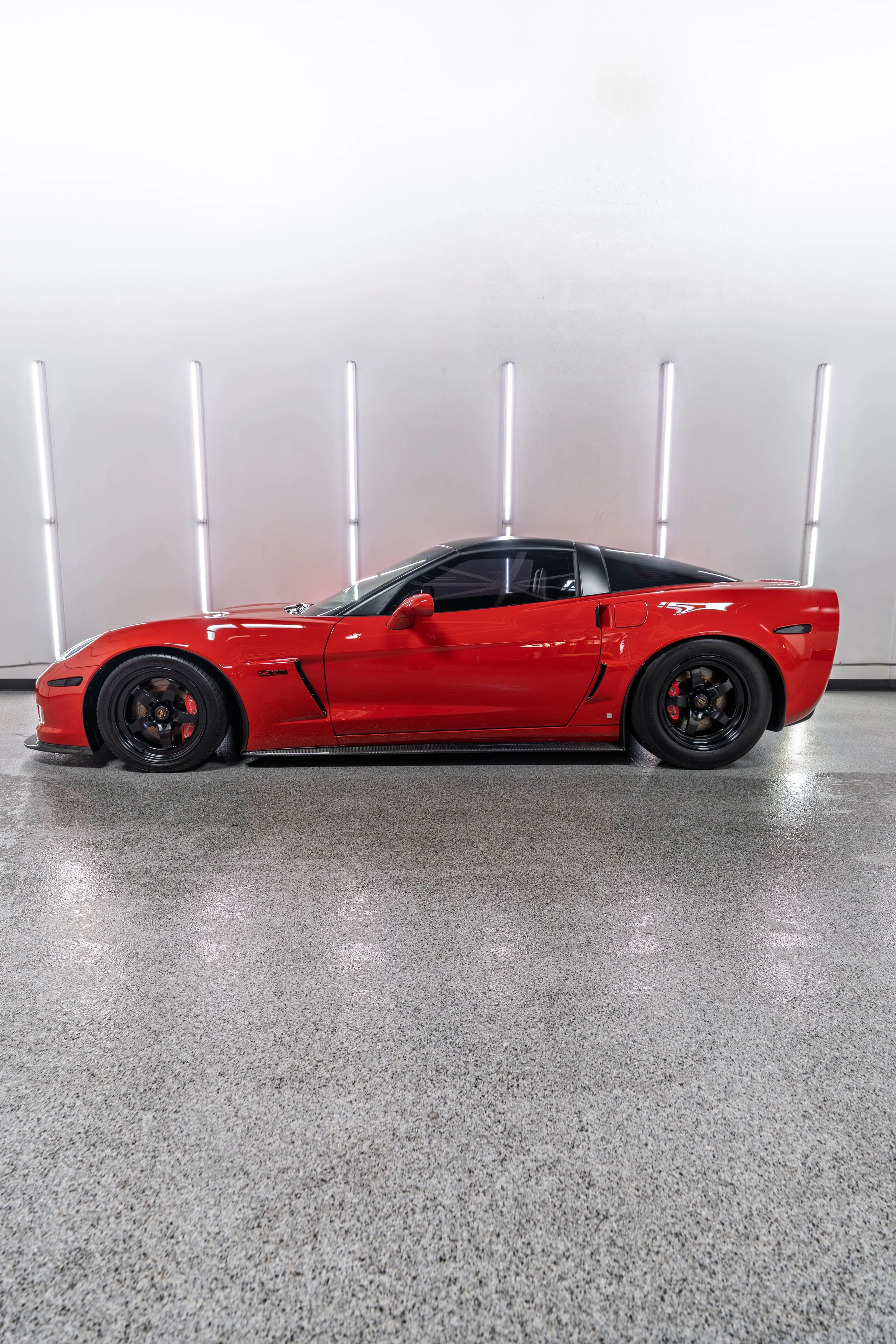 Red sports car parked indoors against a white wall with vertical fluorescent lights.