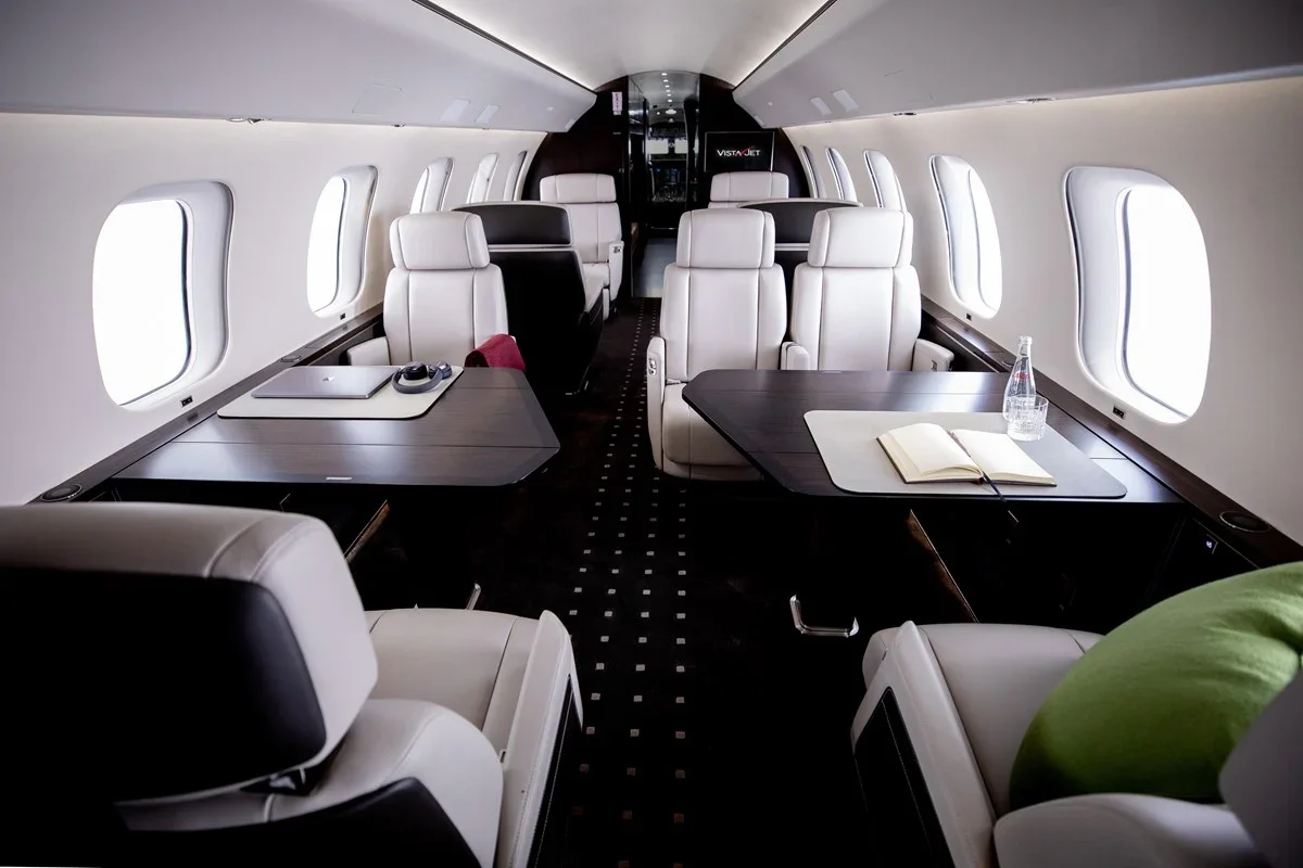 Interior of a private jet with white leather seats, dark wood tables, and windows along the sides.
