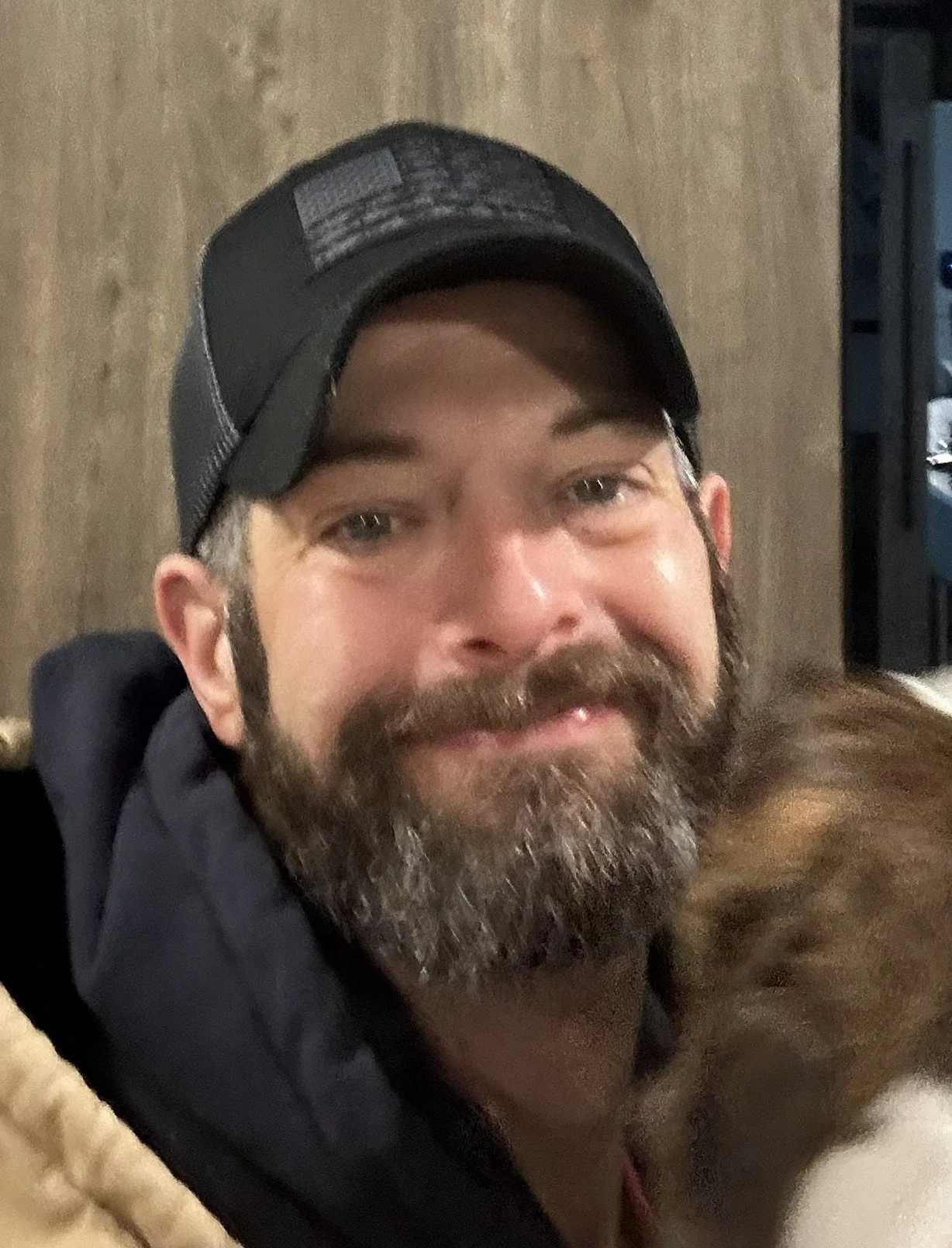 A man with a beard and mustache wearing a black baseball cap, smiling, with a background of wooden wall and some electronic equipment.