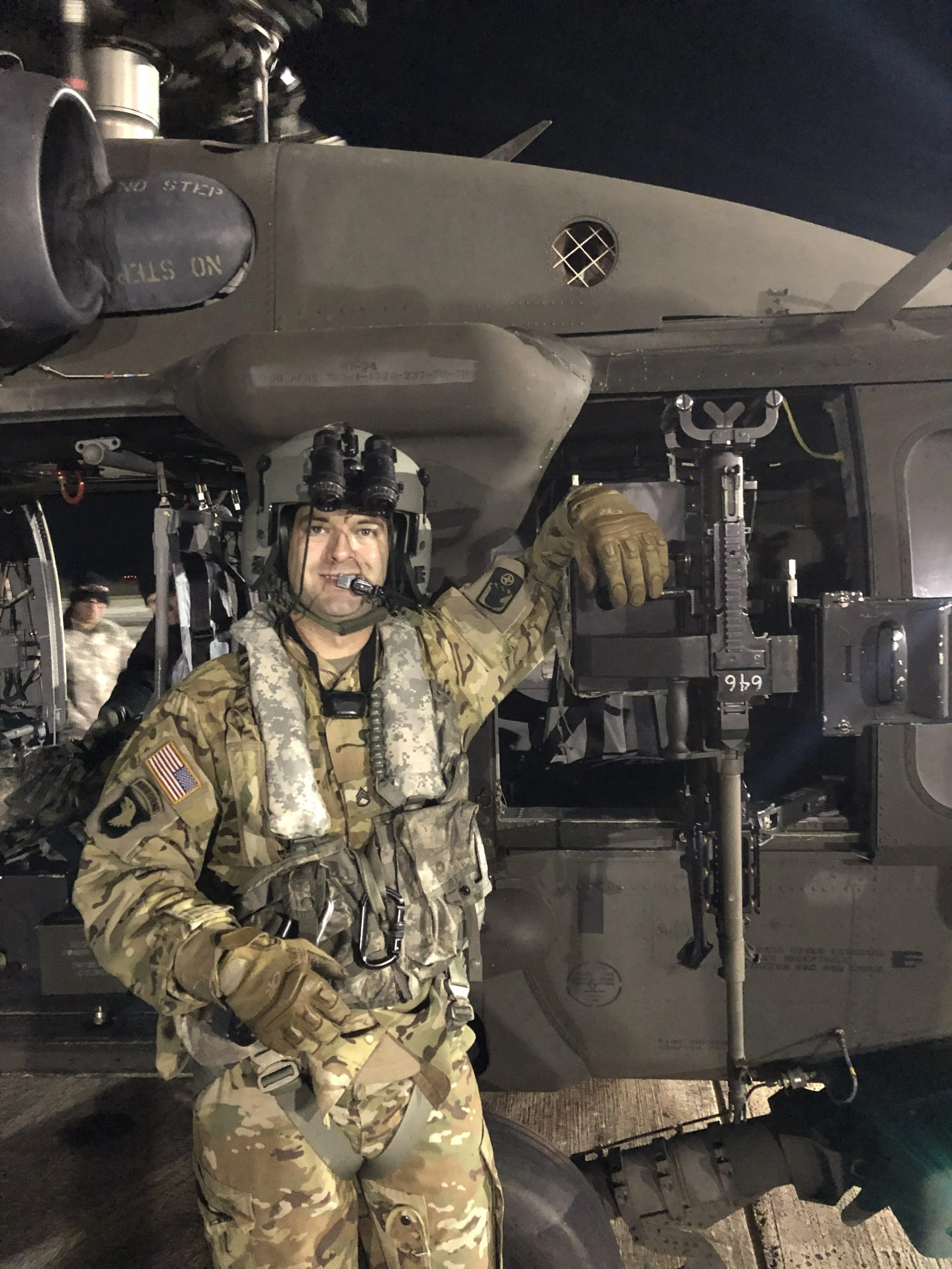 A soldier in military uniform and tactical gear standing beside a military vehicle at night, smiling and pointing with his right hand.