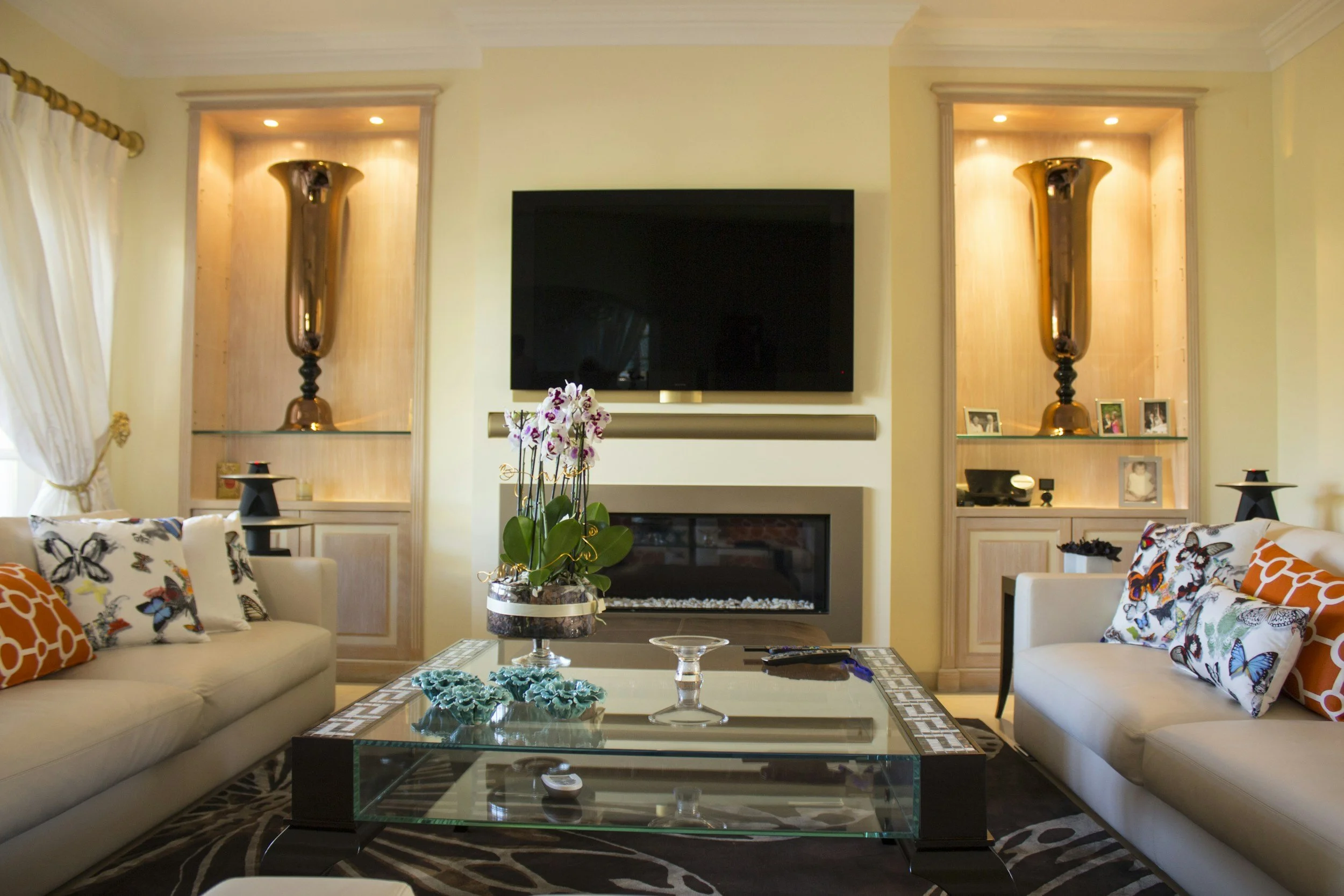 Living room with beige sofas, colorful butterfly and orange patterned pillows, glass coffee table, and built-in wall shelves with large metallic vases, framed photos, and decorative items, TV mounted above a fireplace, orchid on table, and curtains on left window.