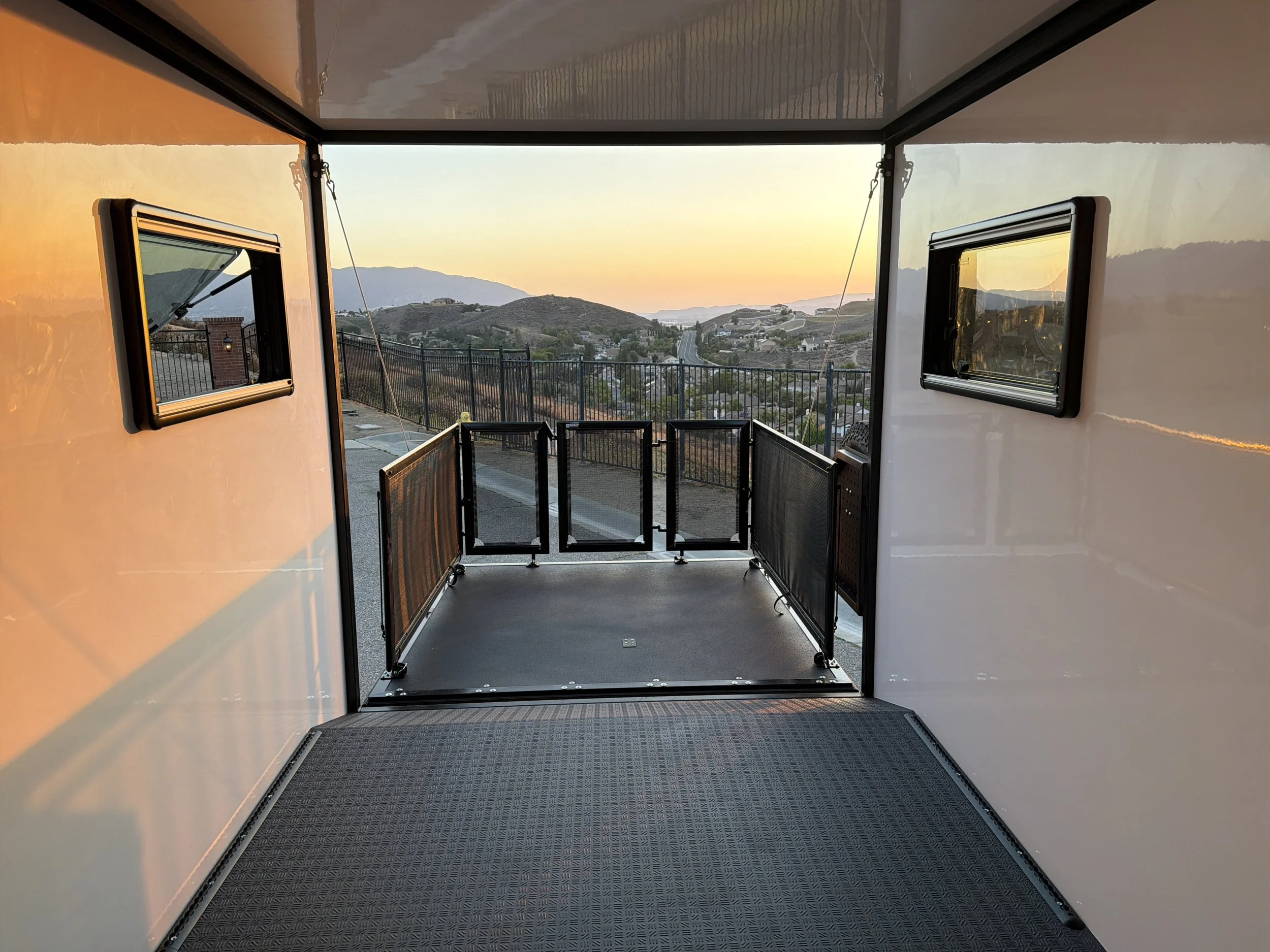 View from inside a transport vehicle, showing an open door with a safety gate, overlooking a hilly landscape during sunset.