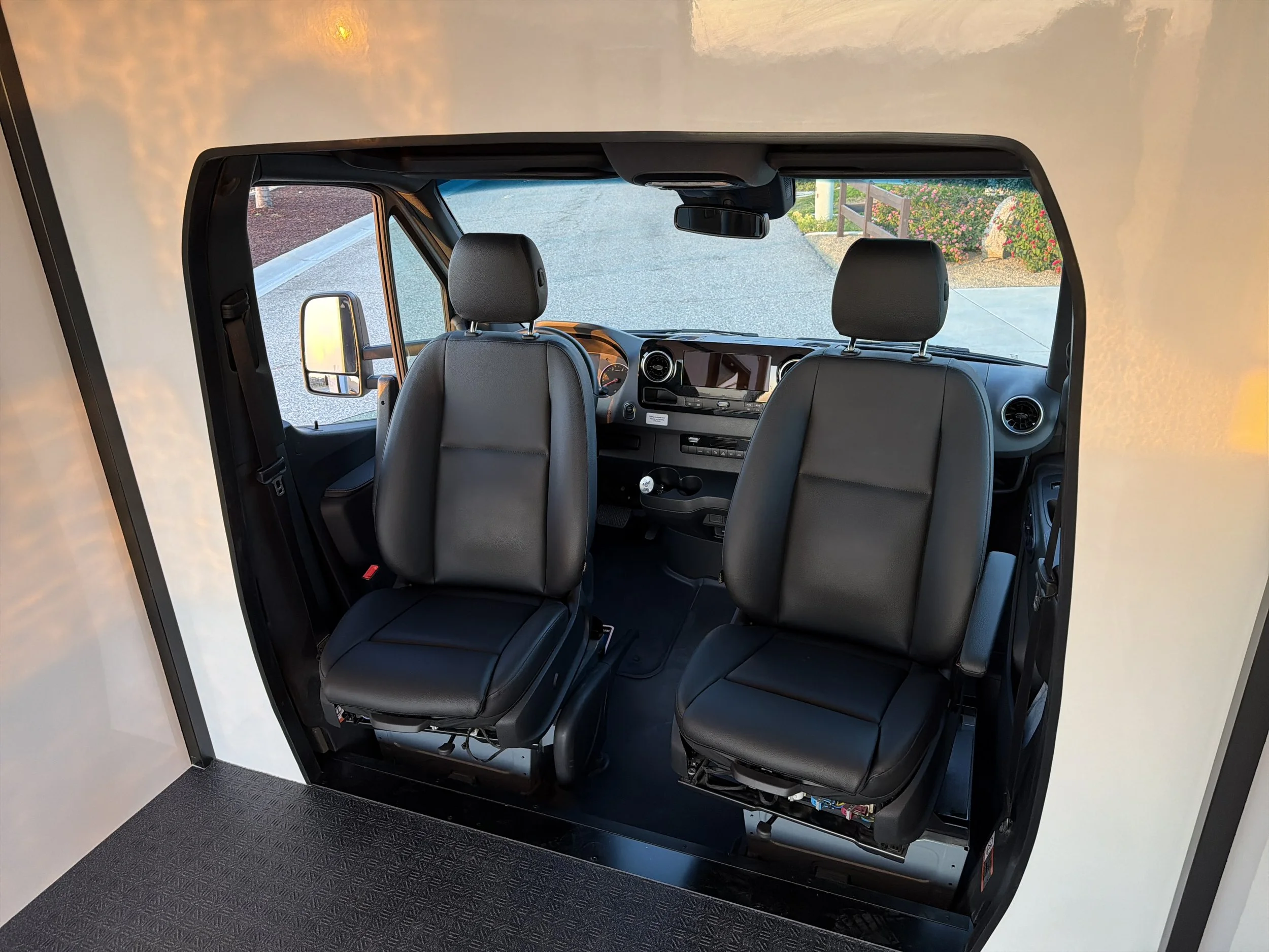The image shows the interior of a vehicle viewed from the passenger side, with two front seats visible, a dashboard, and a windshield. Outside the windshield, there is a paved road, some greenery, and flowering bushes.
