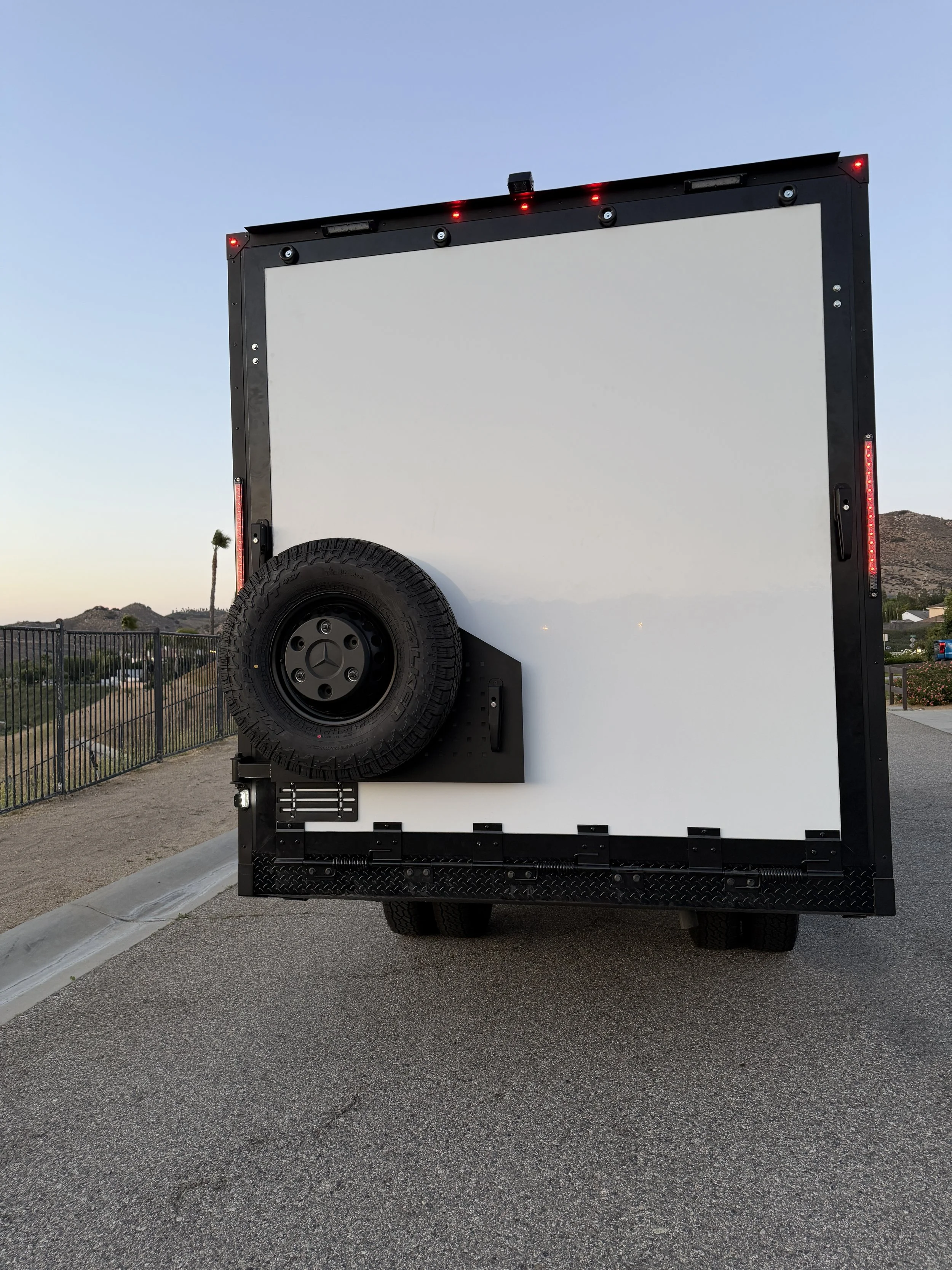 Photo of a mobile movie theater trailer with a large white screen, a spare tire on the side, and red marker lights, parked outdoors near a fence and desert landscape during sunset.