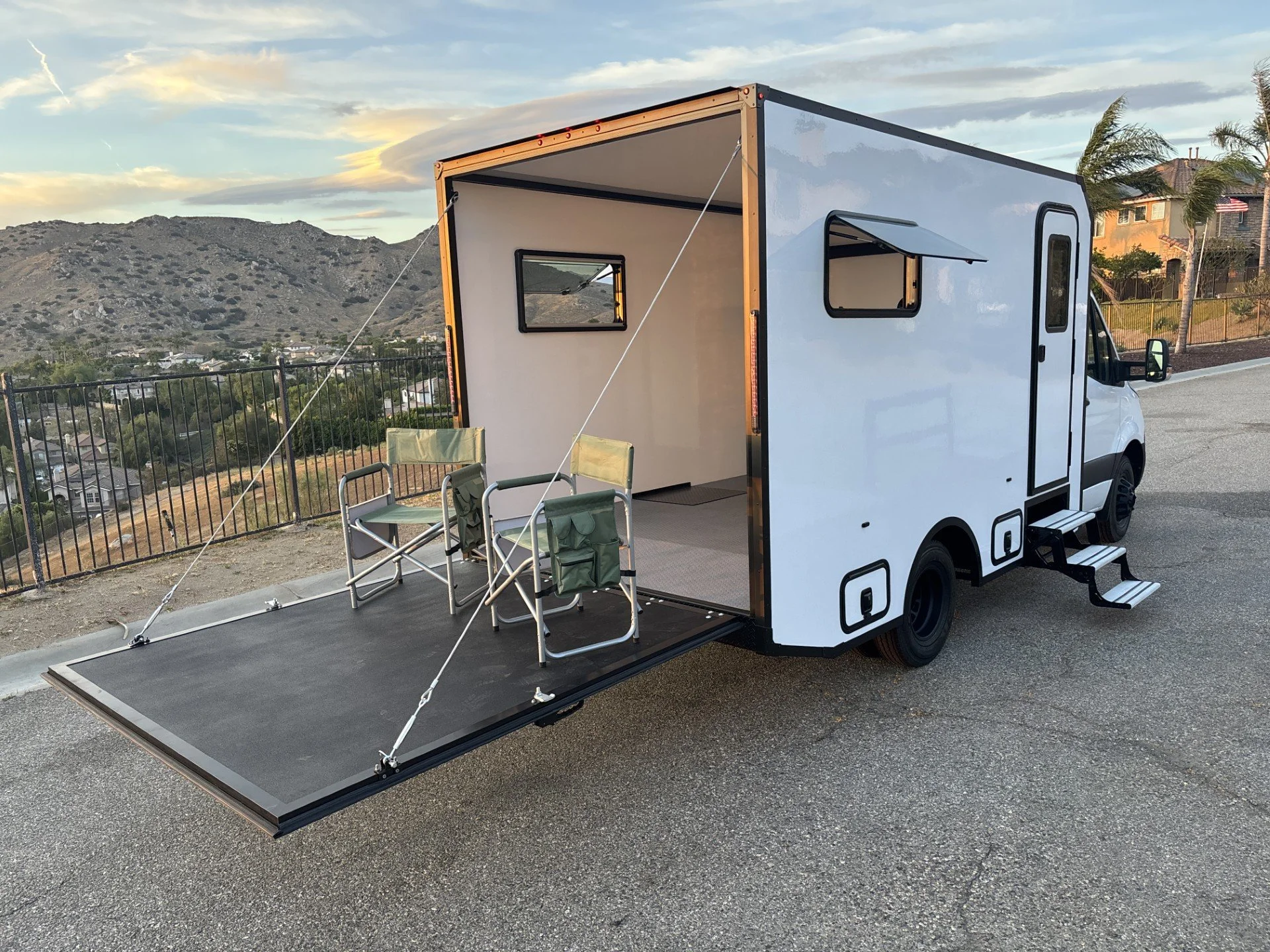 White portable camper vehicle with an extended ramp, two chairs outside, set against a backdrop of hills, houses, and a partly cloudy sky during sunset.
