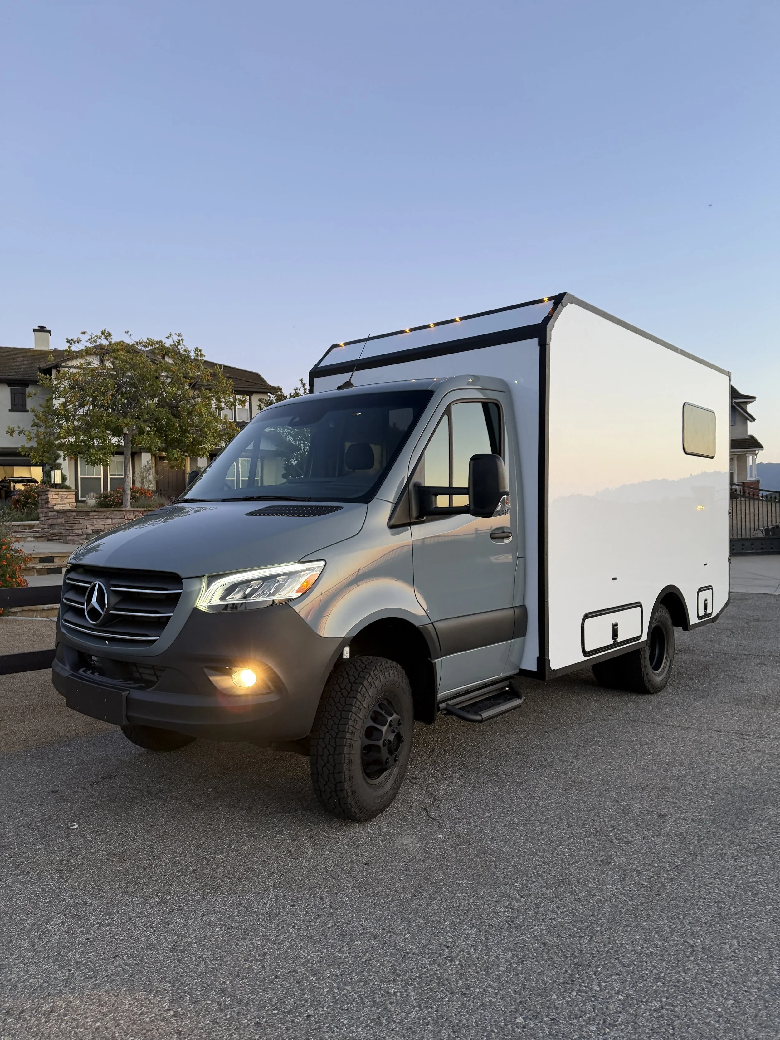 A silver box truck with a Mercedes-Benz logo parked on a residential street during dusk, with houses, trees, and a mountain in the background.