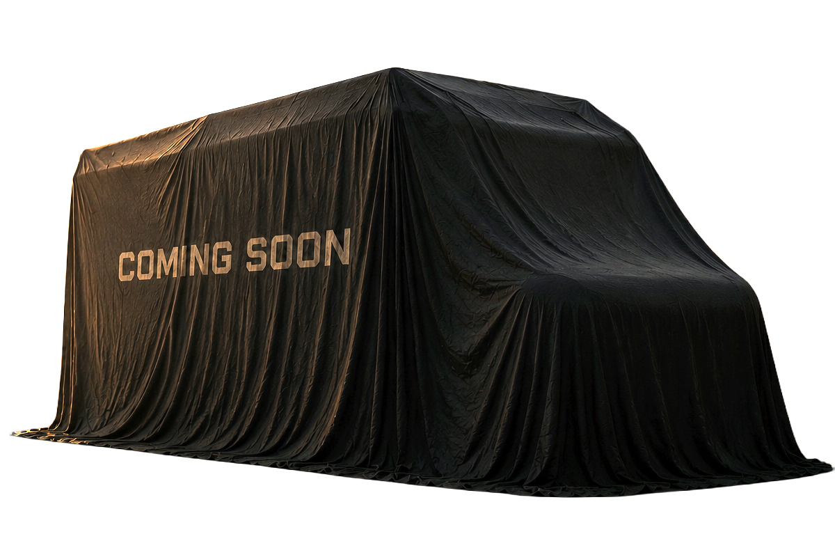 A small black van with a cover that says 'Coming Soon' parked on a sandy beach at sunset with rocky formations and trees in the background.