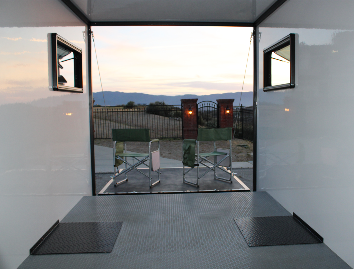 View from inside a trailer looking out onto an outdoor patio with two green chairs, a black metal gate, brick pillars with lights, a dirt pathway, and distant mountains at sunset.