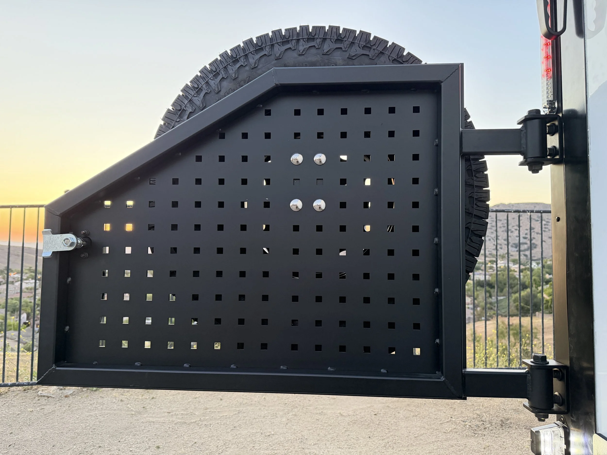 A black metal frame with a partial spare tire mounted on it and a battery attached to the side, outdoors during sunset or sunrise with a clear sky and distant hills or mountains.