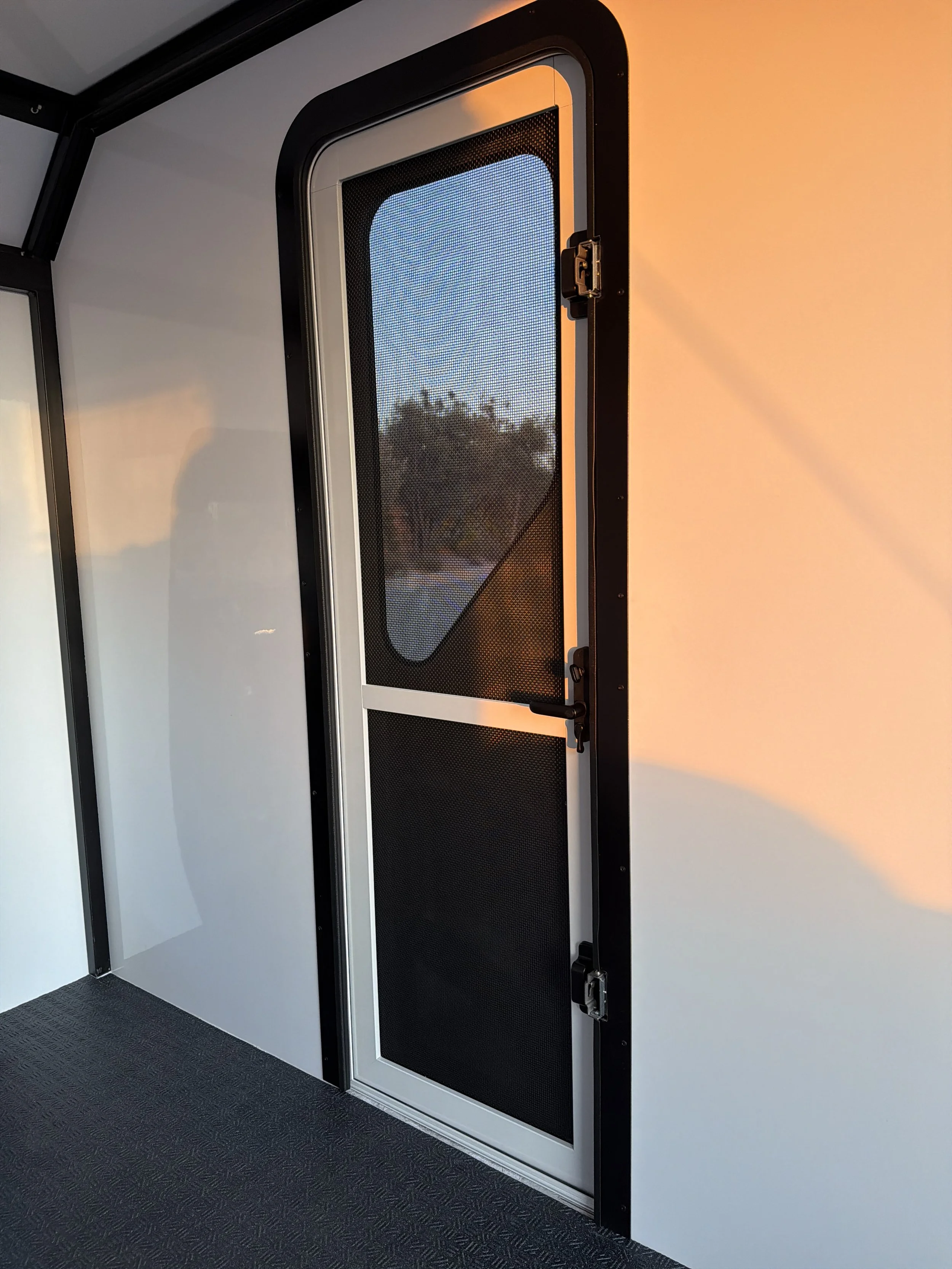 A black metal door with a screen window and latch, set in a white wall, with sunlight casting shadow on the wall.