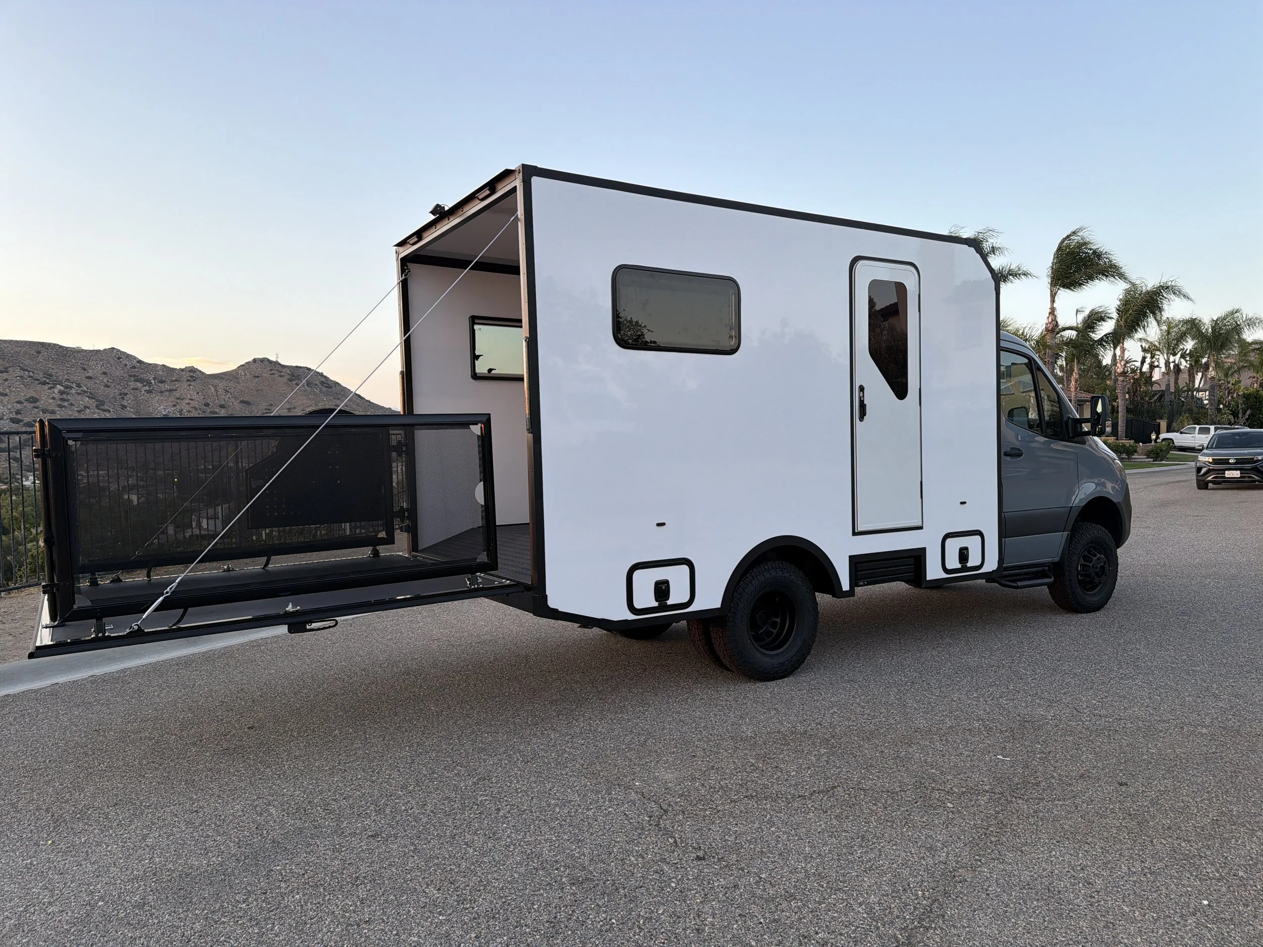 A white camper van parked on a paved road with a black cargo area at the back, in a scenic area with mountains and palm trees in the background during sunset or sunrise.