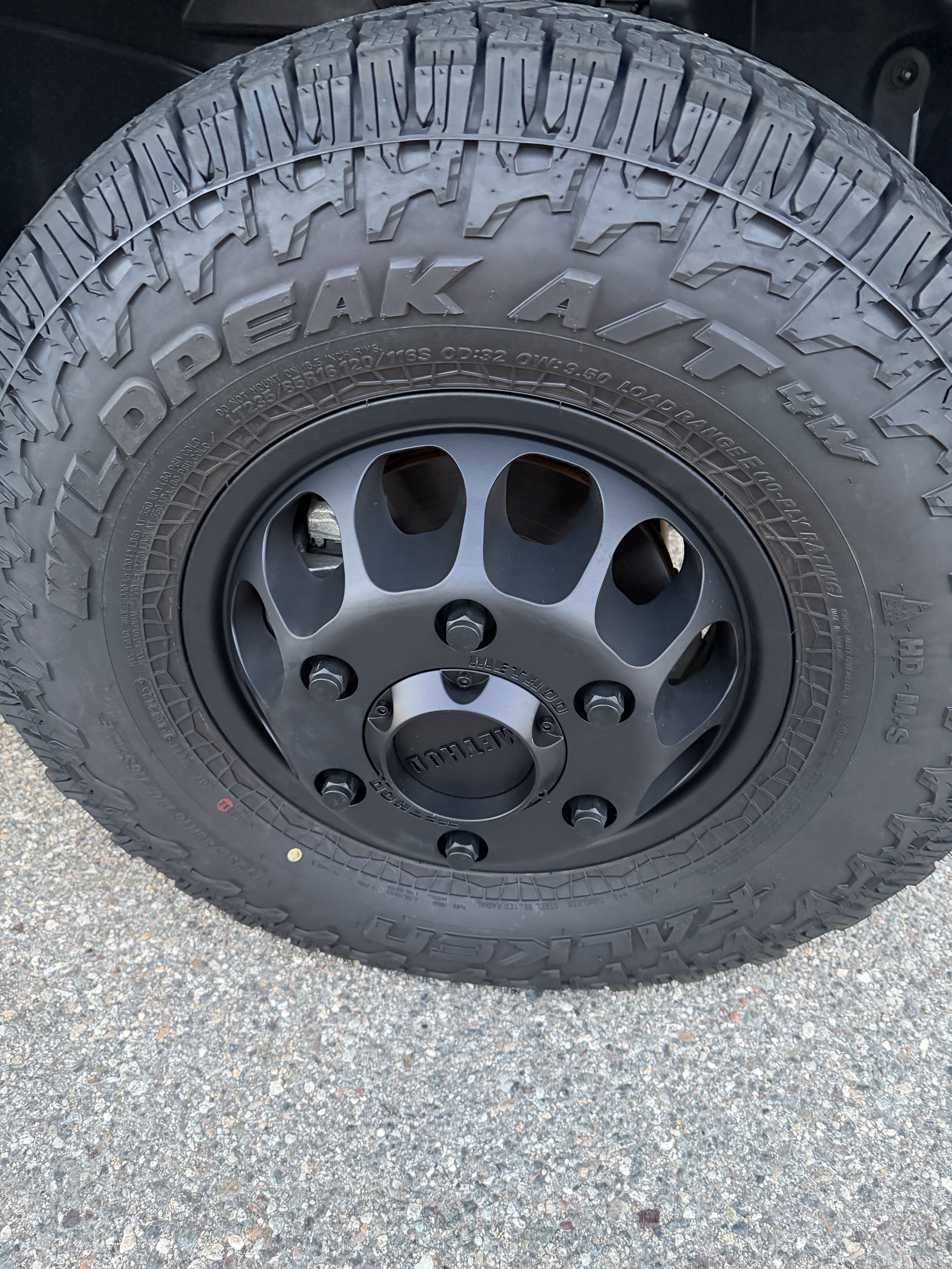 Close-up of a black tire with large rugged tread pattern and a matte black wheel with circular holes, mounted on a vehicle, on a gravel surface.