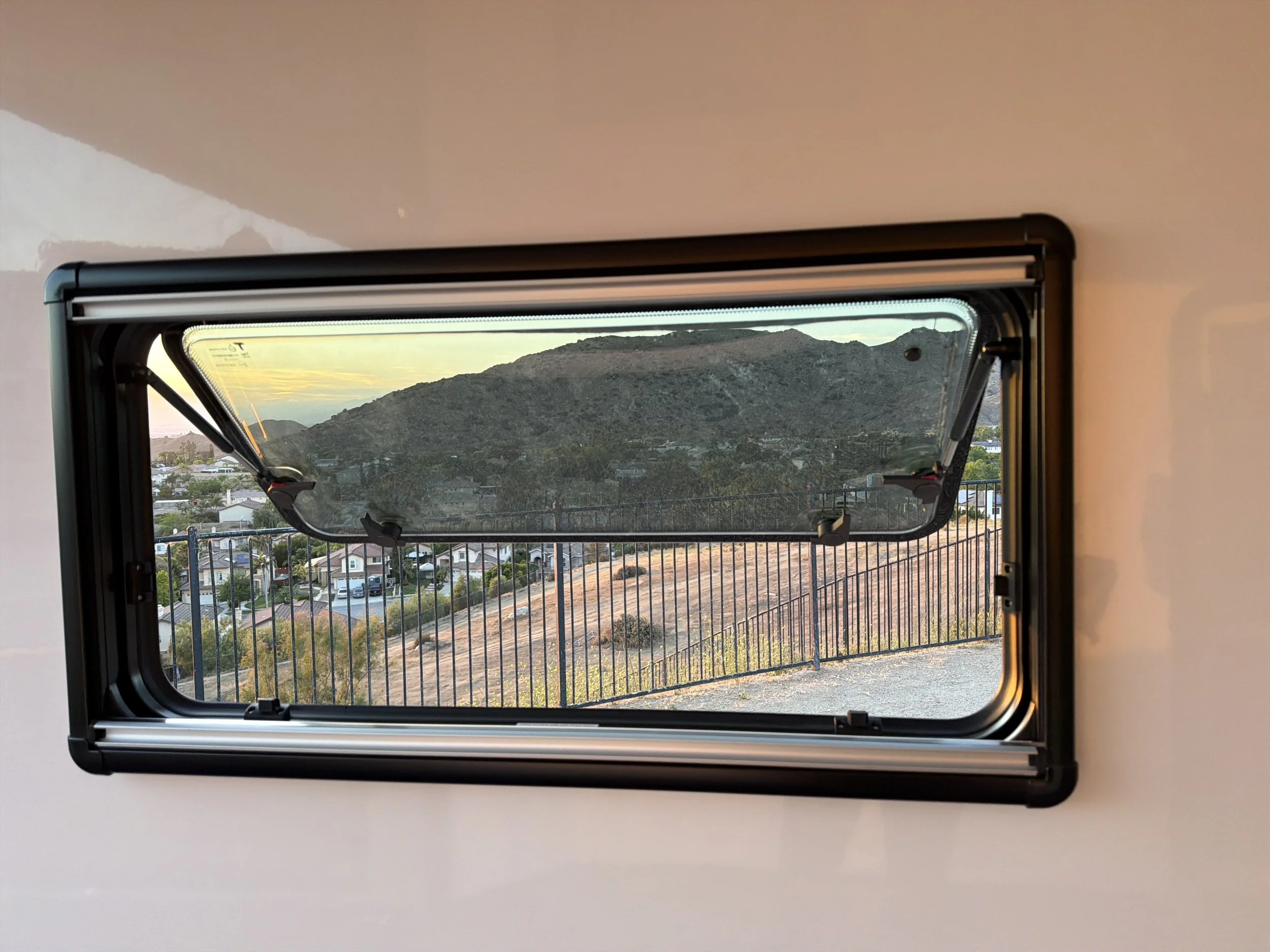A landscape view of hills and houses through a partially open window with a black frame and a screen on the bottom half, set in a room with beige walls.
