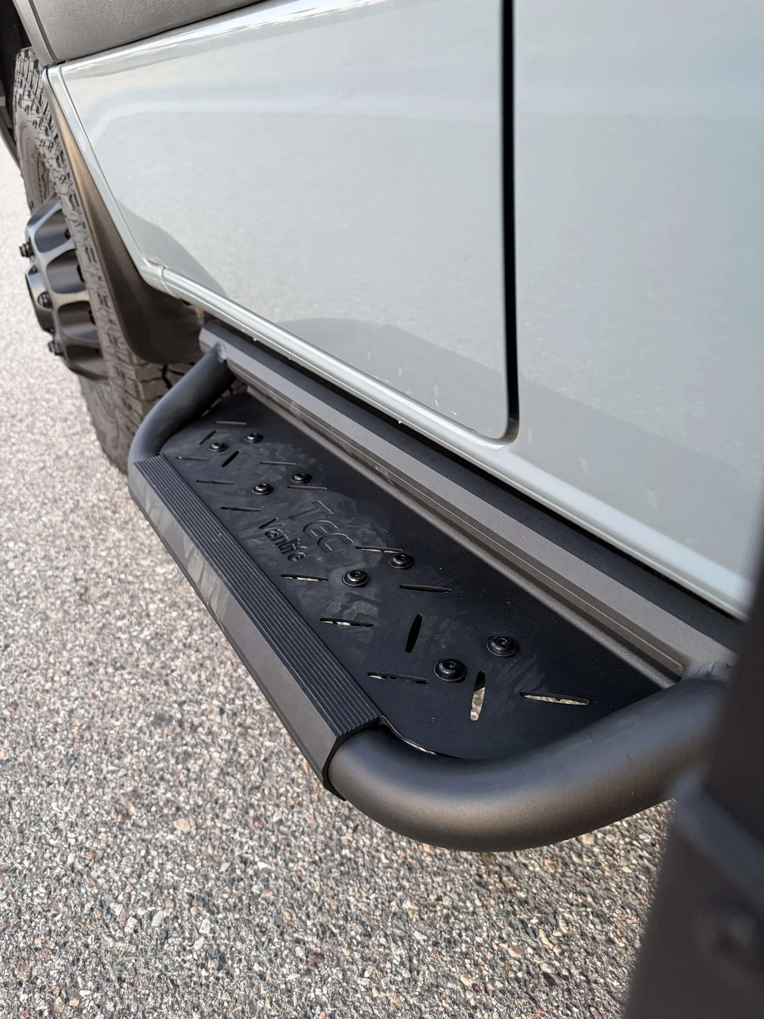 Close-up of the side door and wheel of a silver SUV, with a black plastic step below and part of the gravel ground visible underneath.