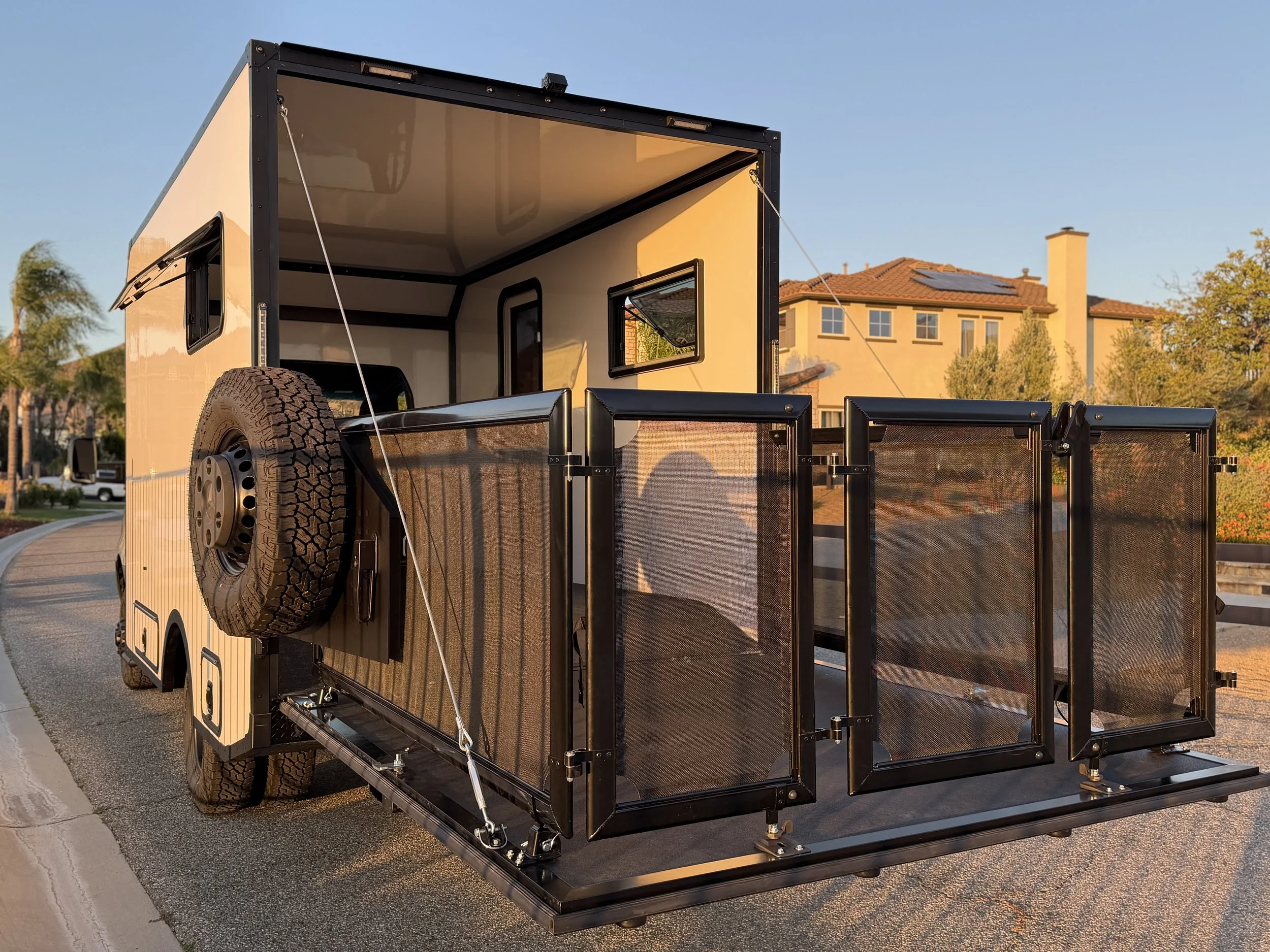 A portable trailer with an open ramp door, a tire mounted on its side, and window panels, parked on a residential street during sunset.