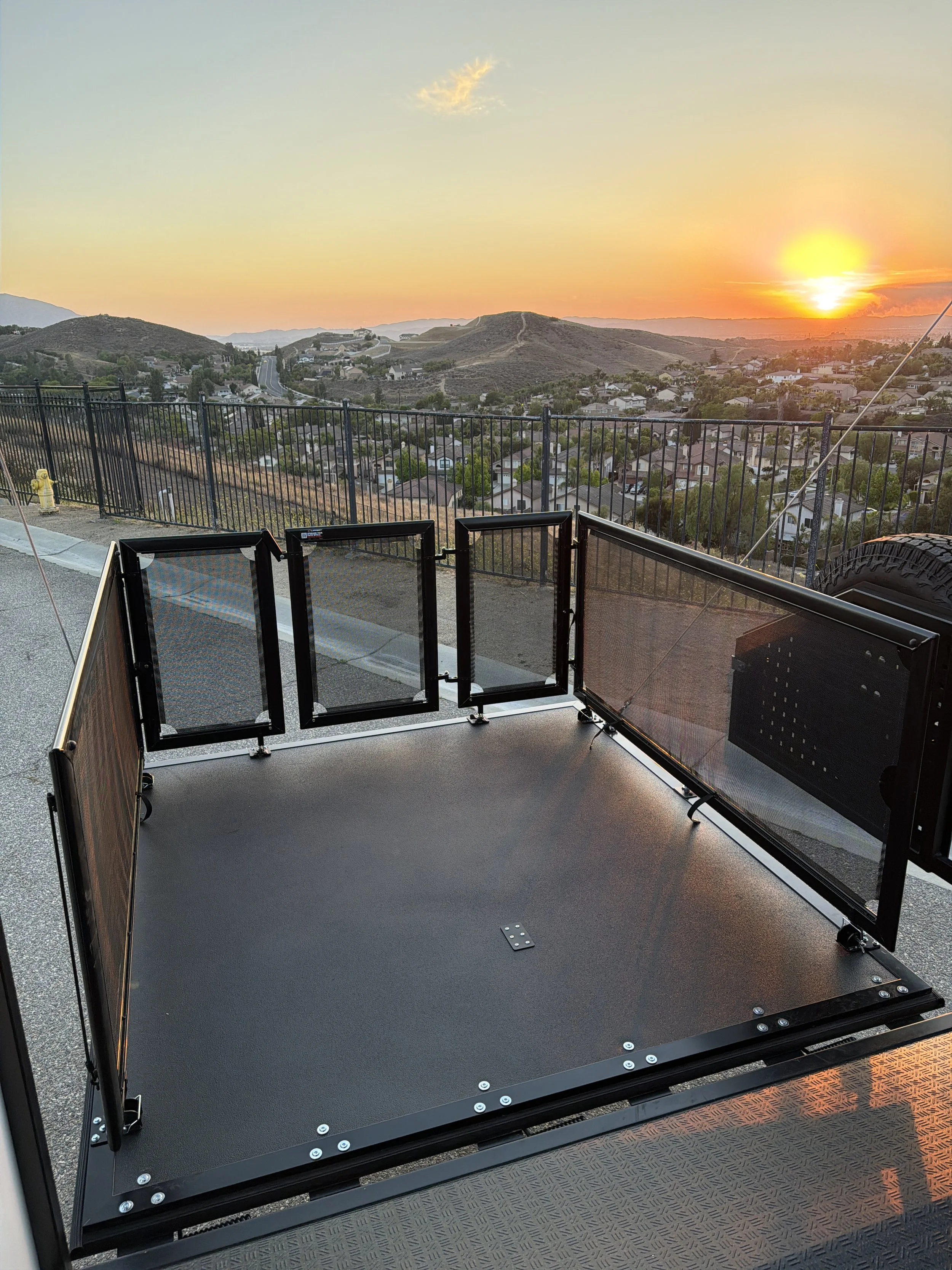 A view from a rooftop or balcony during sunset, showing houses and rolling hills in the distance, with a black metal safety railing and a partially open glass panel in the foreground.