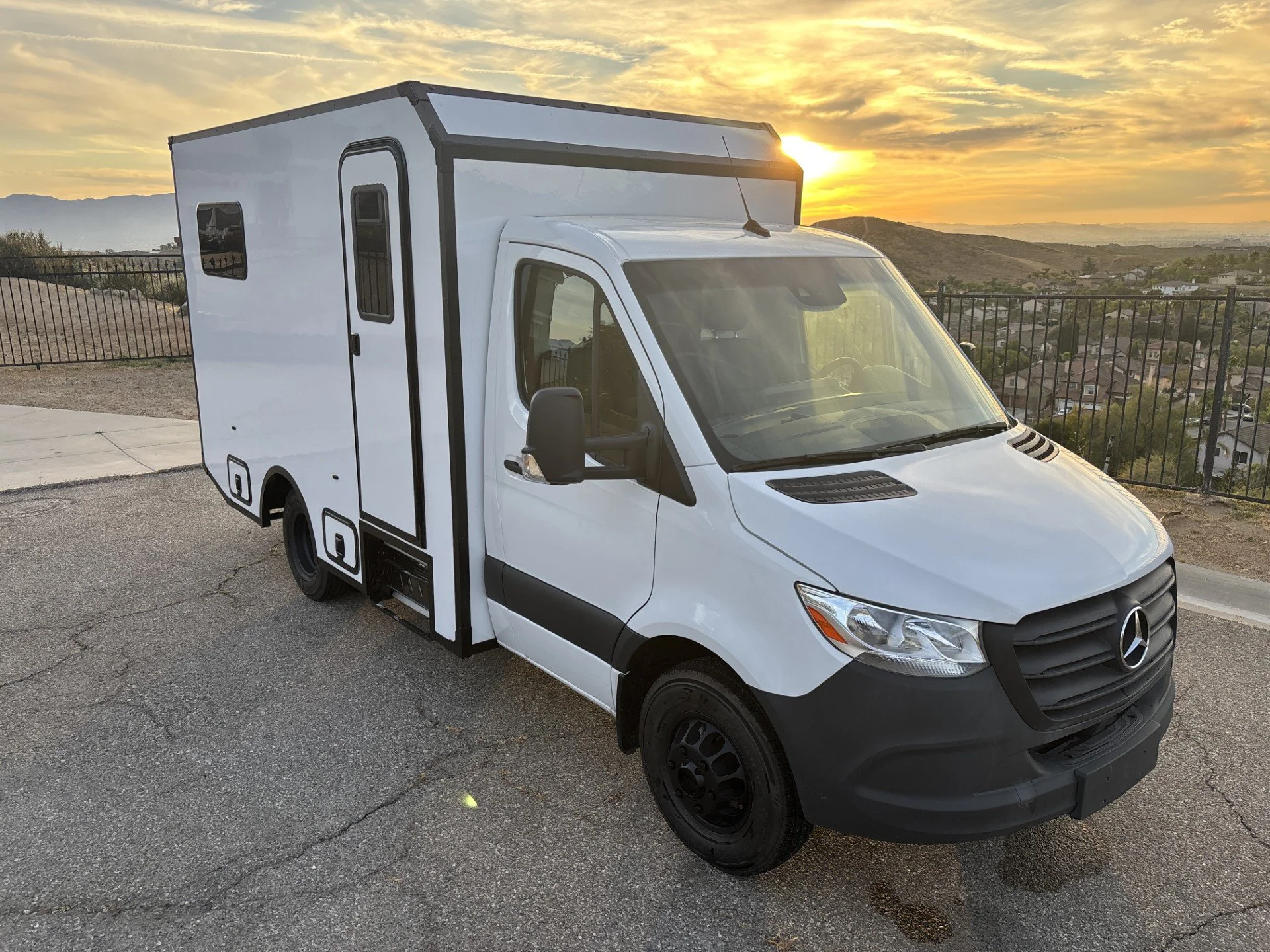 White camper van parked on cracked asphalt with a sunset sky, mountains, and a residential neighborhood in the background.