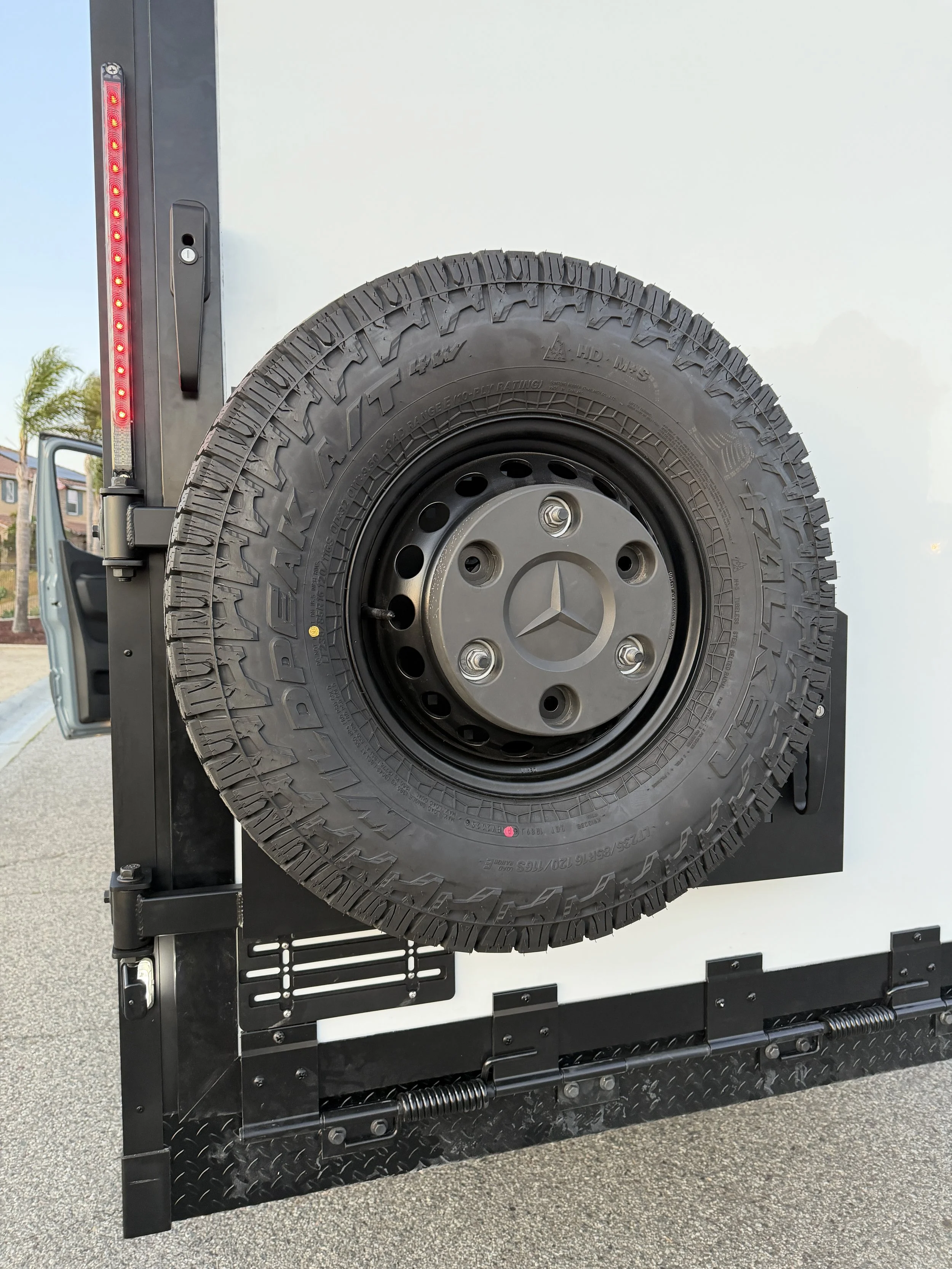 Close-up of a spare tire with a tire cover featuring a Mercedes-Benz logo mounted on the side of a vehicle, with part of the vehicle's black frame and a red taillight visible.
