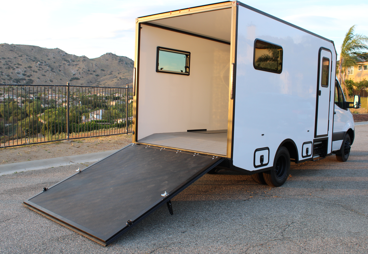 A white box truck with an open back, featuring a ramp leading up into the cargo area. The truck is parked on a street with a fence and hills in the background.