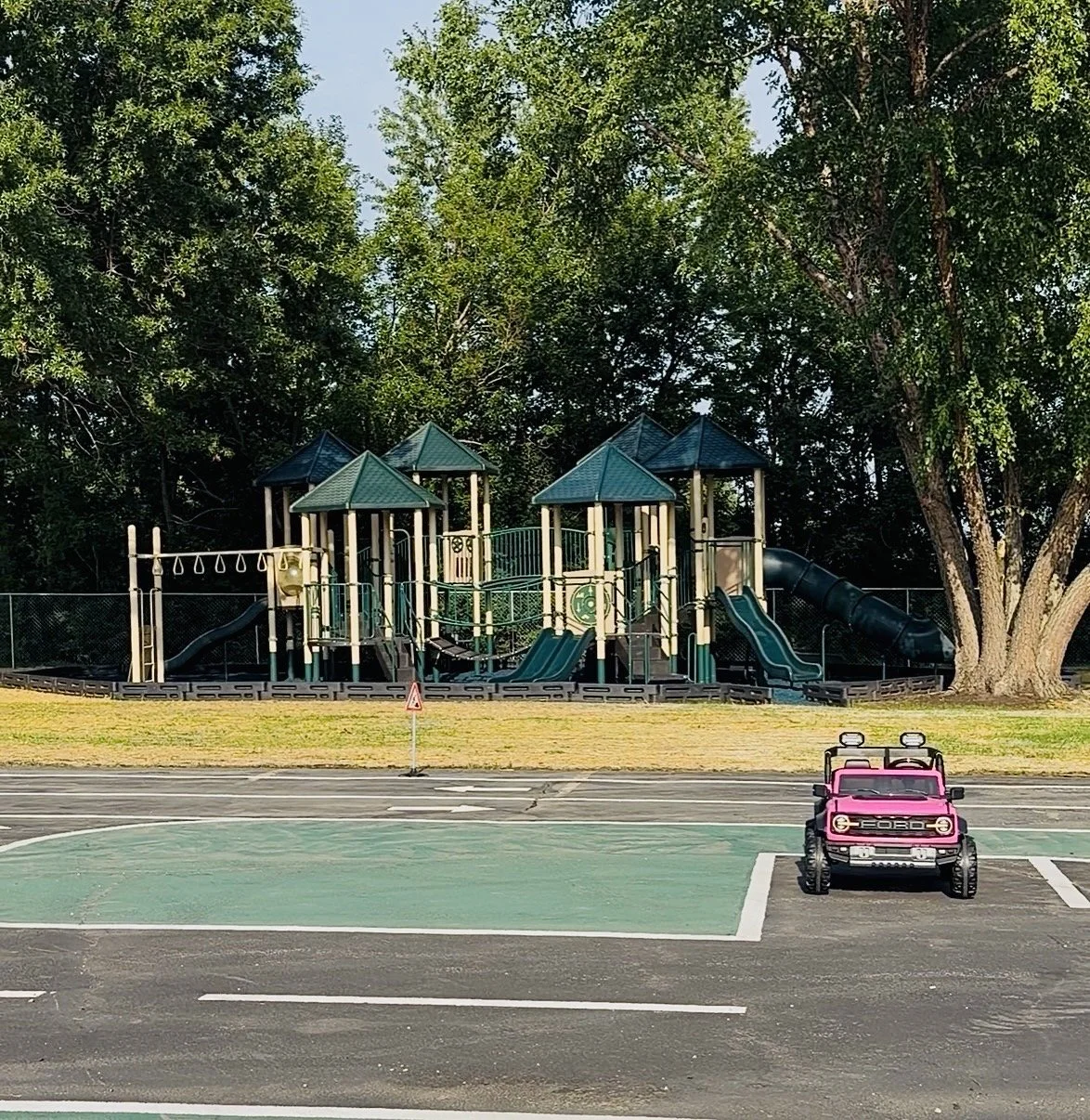 Empty outdoor playground with slides, bridges, and climbing structures, surrounded by trees. A children's pink ride-on toy car is parked nearby on a paved area.