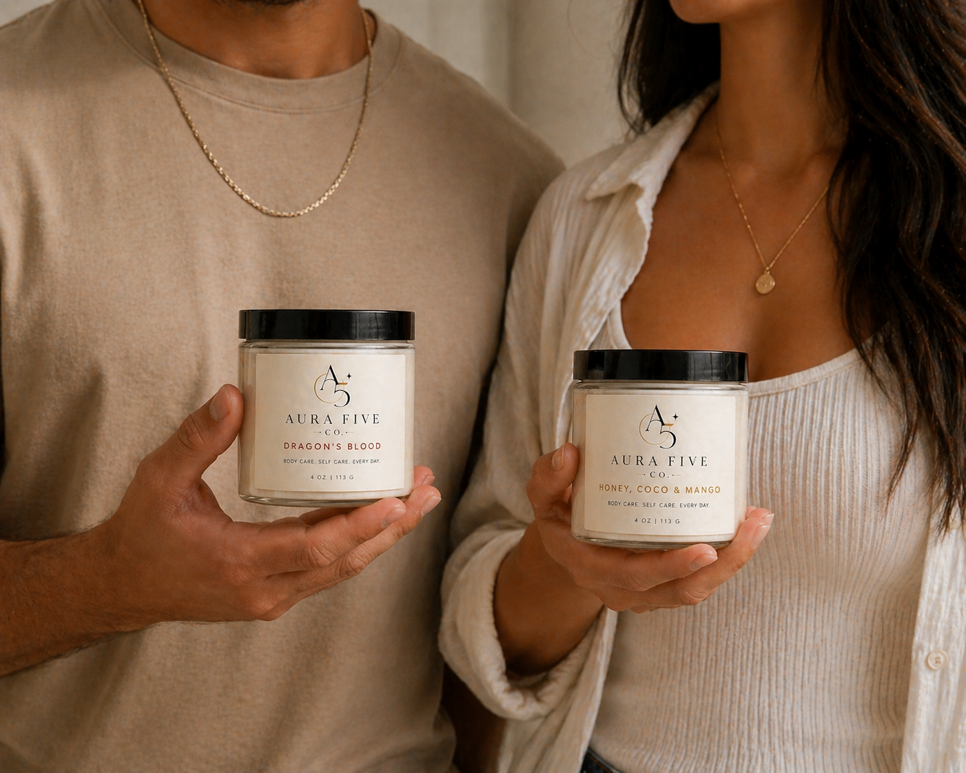 Two people holding jars of Aura Five body care products, one labeled 'Dragon's Blood' and the other 'Honey, Coco & Mango.'