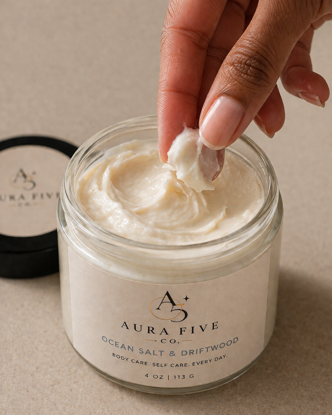 A person taking a scoop of creamy body butter from an open jar labeled Aura Five Co. Ocean Salt & Driftwood.