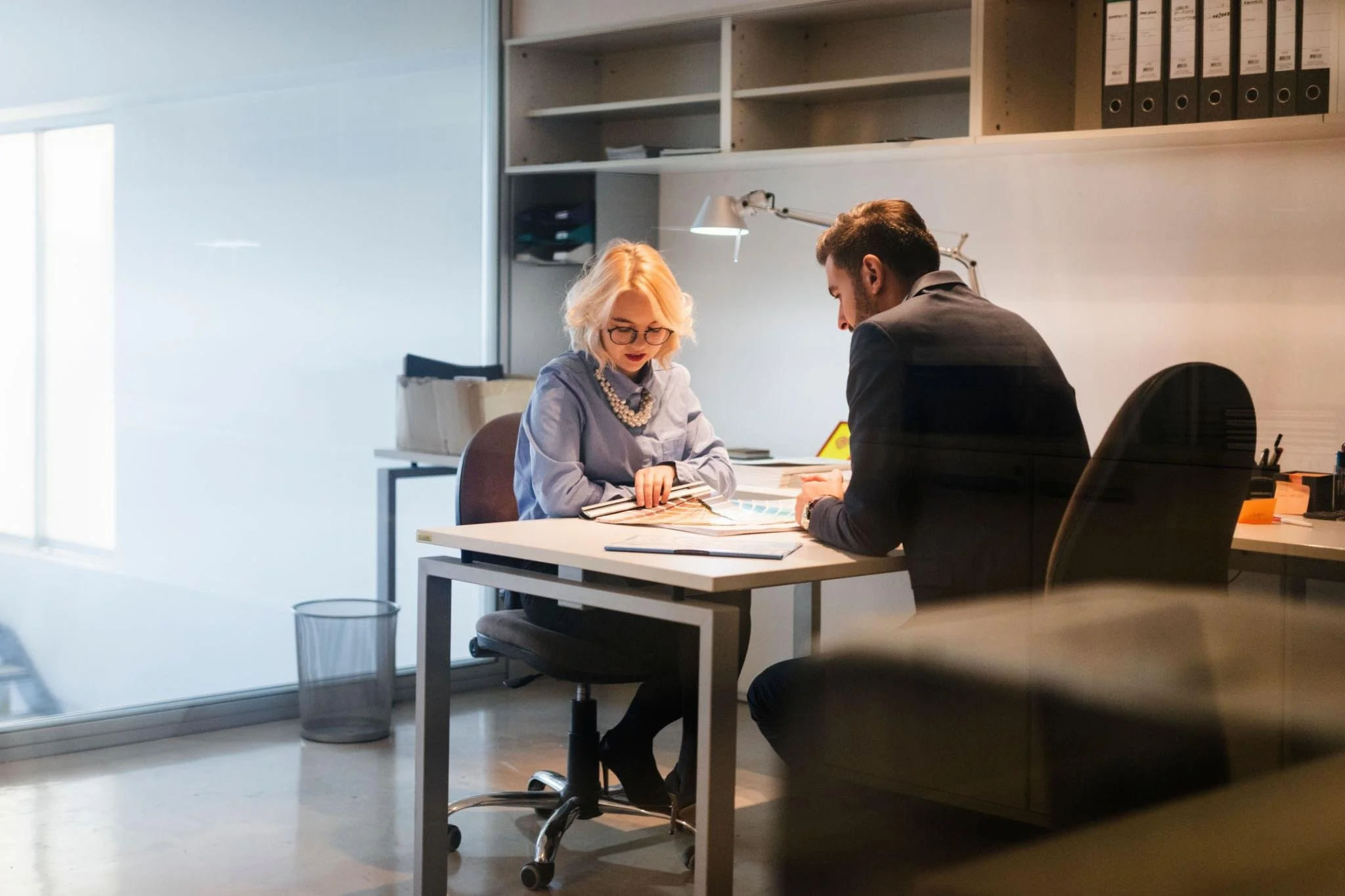 Two colleagues, a woman with blonde hair and glasses and a man with dark hair, sitting at a white desk with a color sample book, discussing work in a modern office. Shelves with binders and folders are in the background.