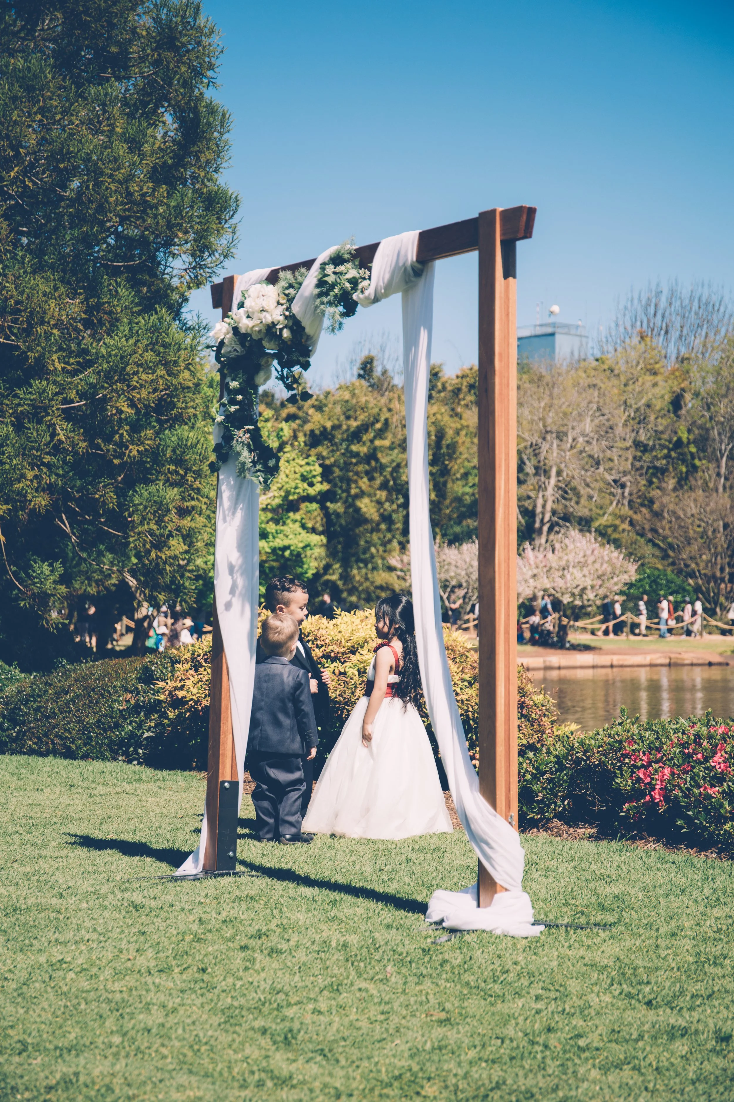 A wedding ceremony takes place outdoors on a sunny day, with a bride and groom standing under a wooden arch decorated with white fabric and flowers, surrounded by trees and greenery.