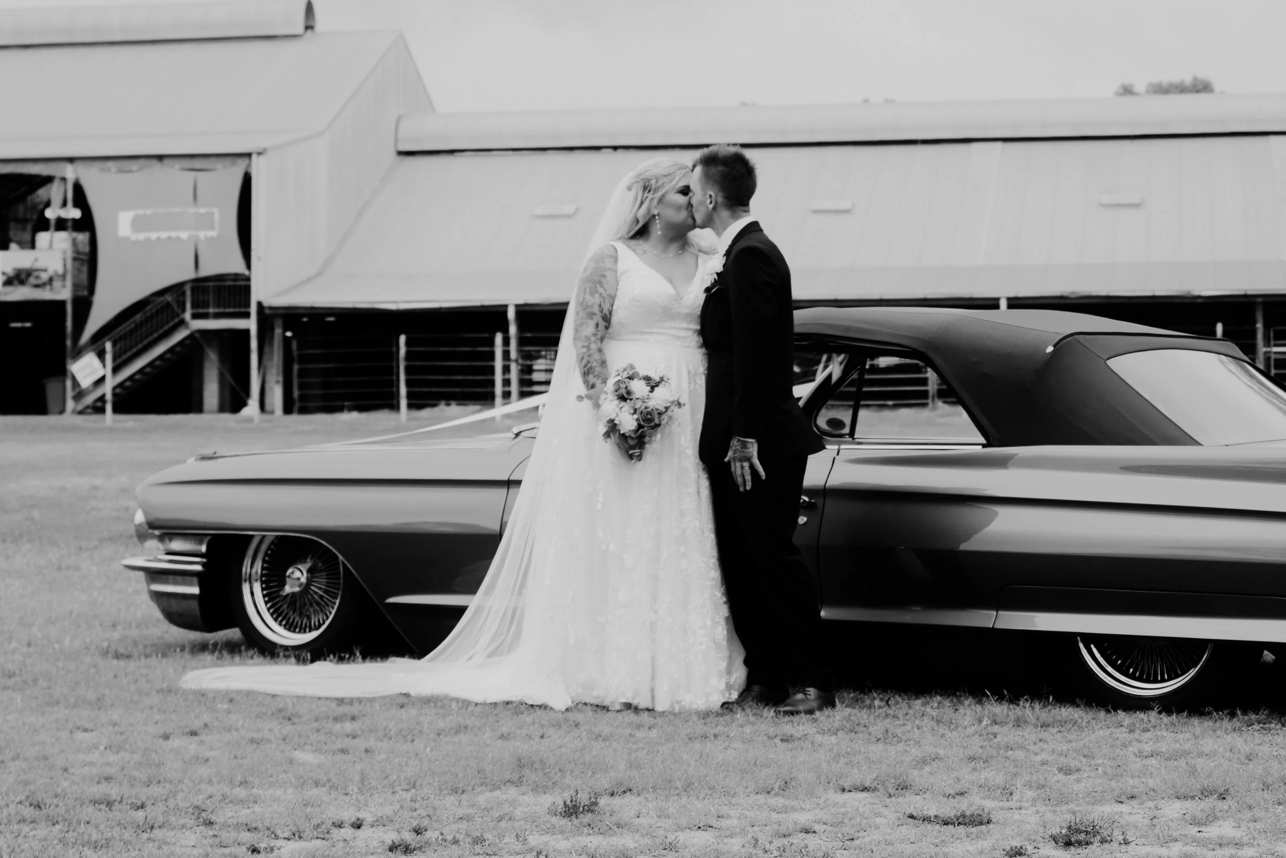 A bride and groom kissing beside a classic car outdoors during daytime with a building in the background.