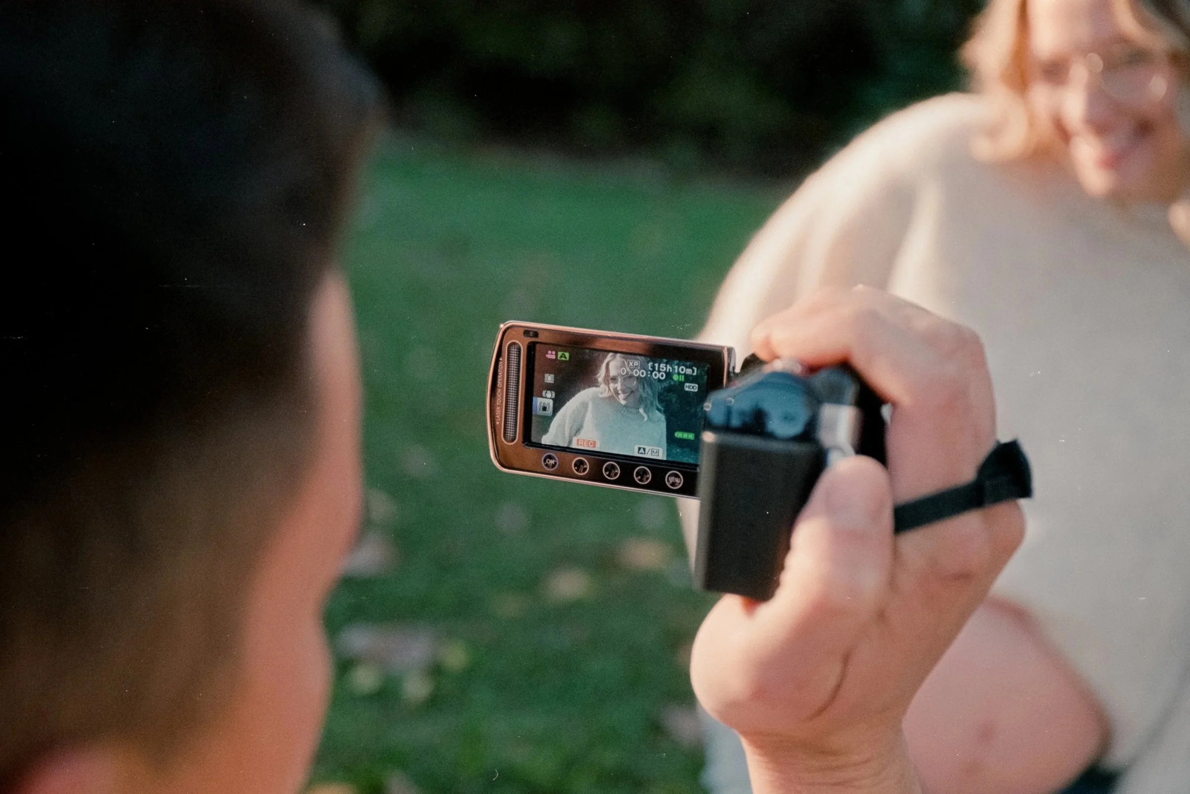 Person filming a woman with a camera in an outdoor setting with green grass.