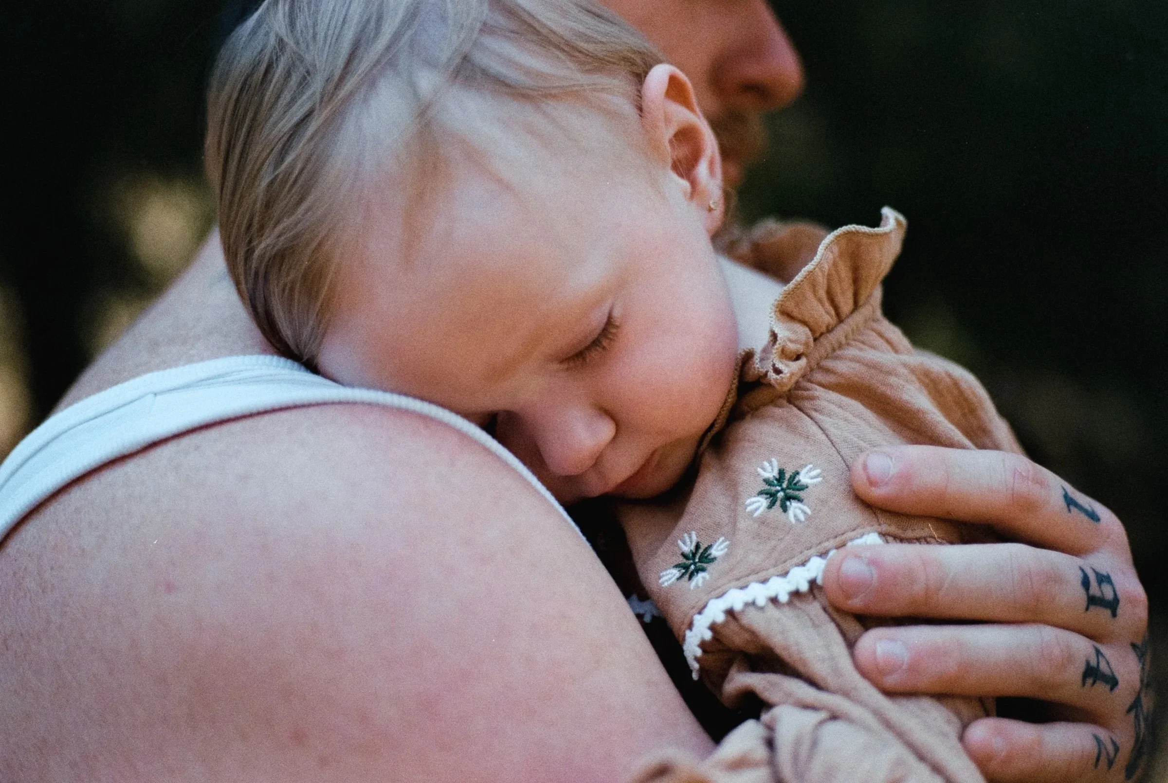 A young child with blonde hair is sleeping peacefully while being embraced by an adult, possibly in an outdoor setting with blurred natural background.