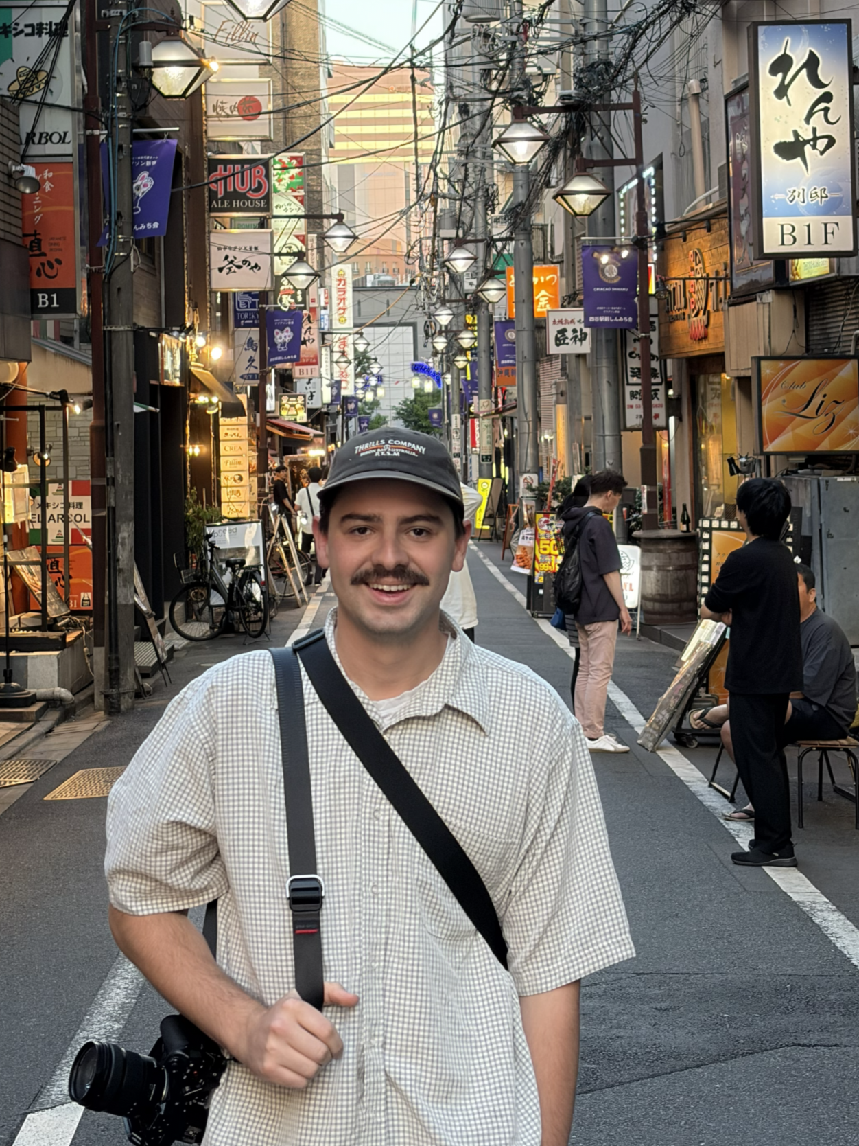 A smiling young man with a mustache wearing a black cap, light-colored checkered shirt, and carrying a camera with a strap and a bag, standing in a busy, narrow city street with signs in Japanese, streetlights, and several pedestrians in the background.