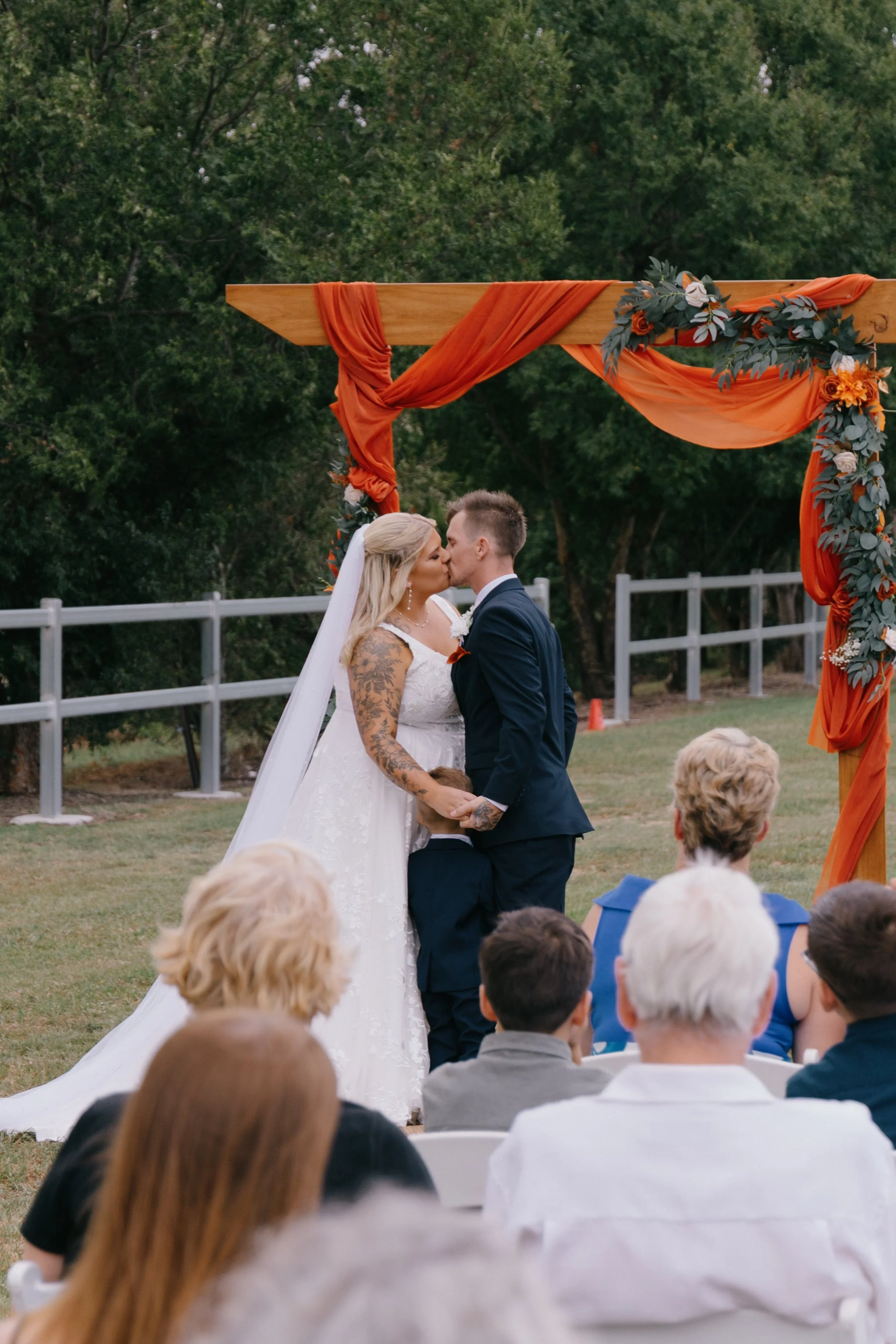 A bride and groom share a kiss during their outdoor wedding ceremony under an orange draped arch decorated with flowers, with guests seated in front of them.