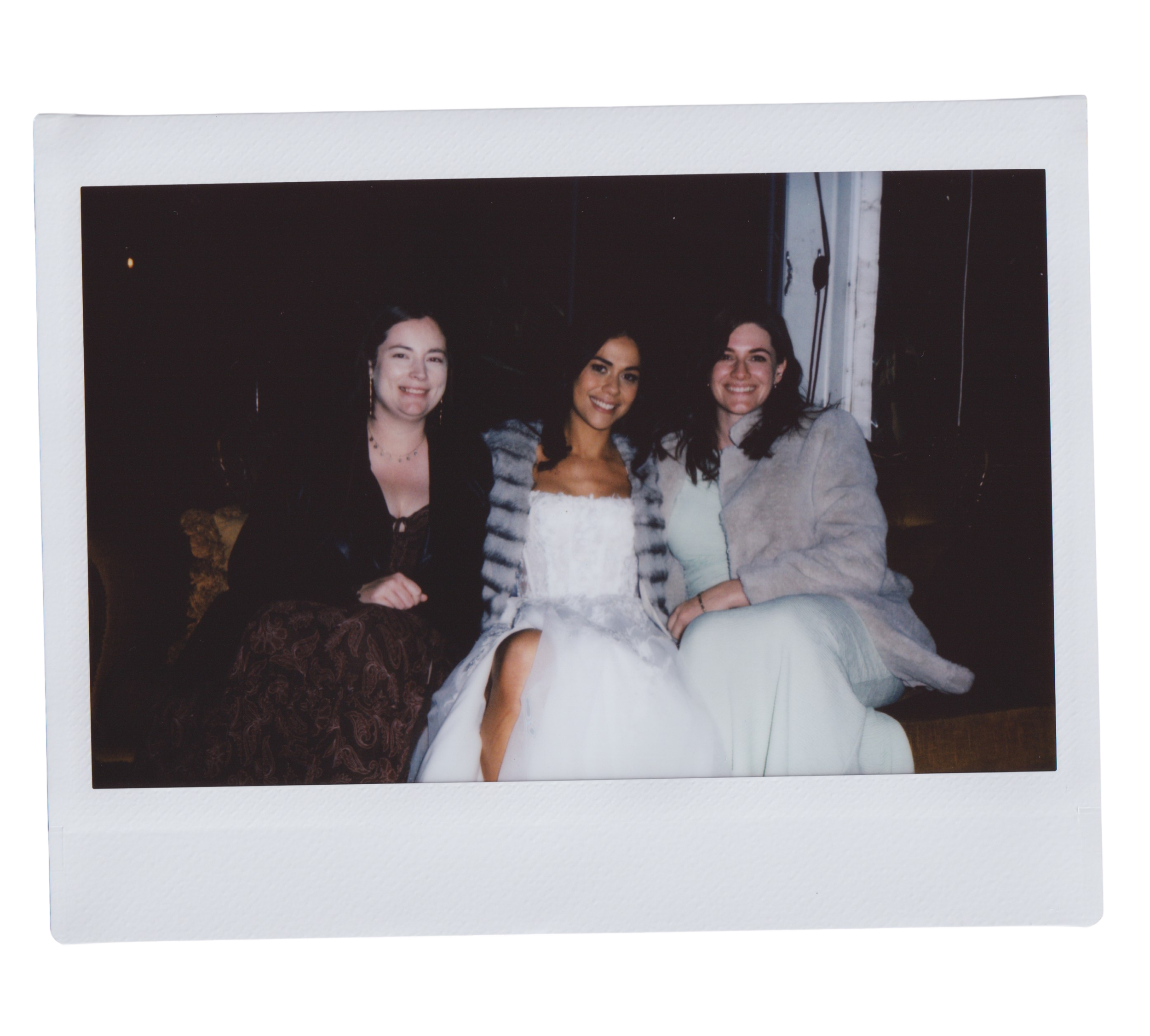 Photograph of three smiling women sitting close together indoors, with dark walls and visible electrical wiring behind them.