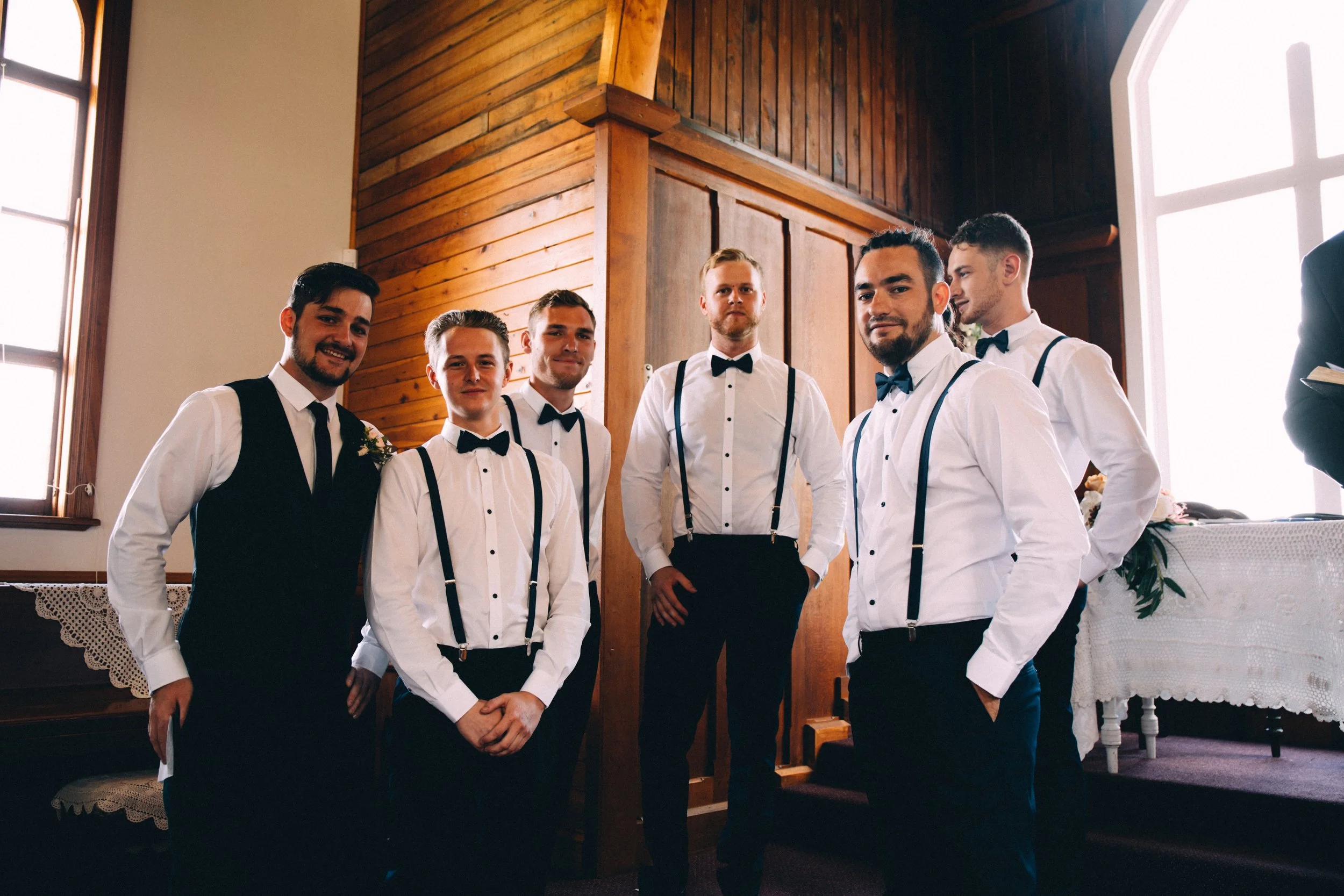 A group of seven men dressed in formal attire with white shirts, black bow ties, and suspenders, standing inside a room with wood-paneled walls and large windows, smiling and posing for a photo.
