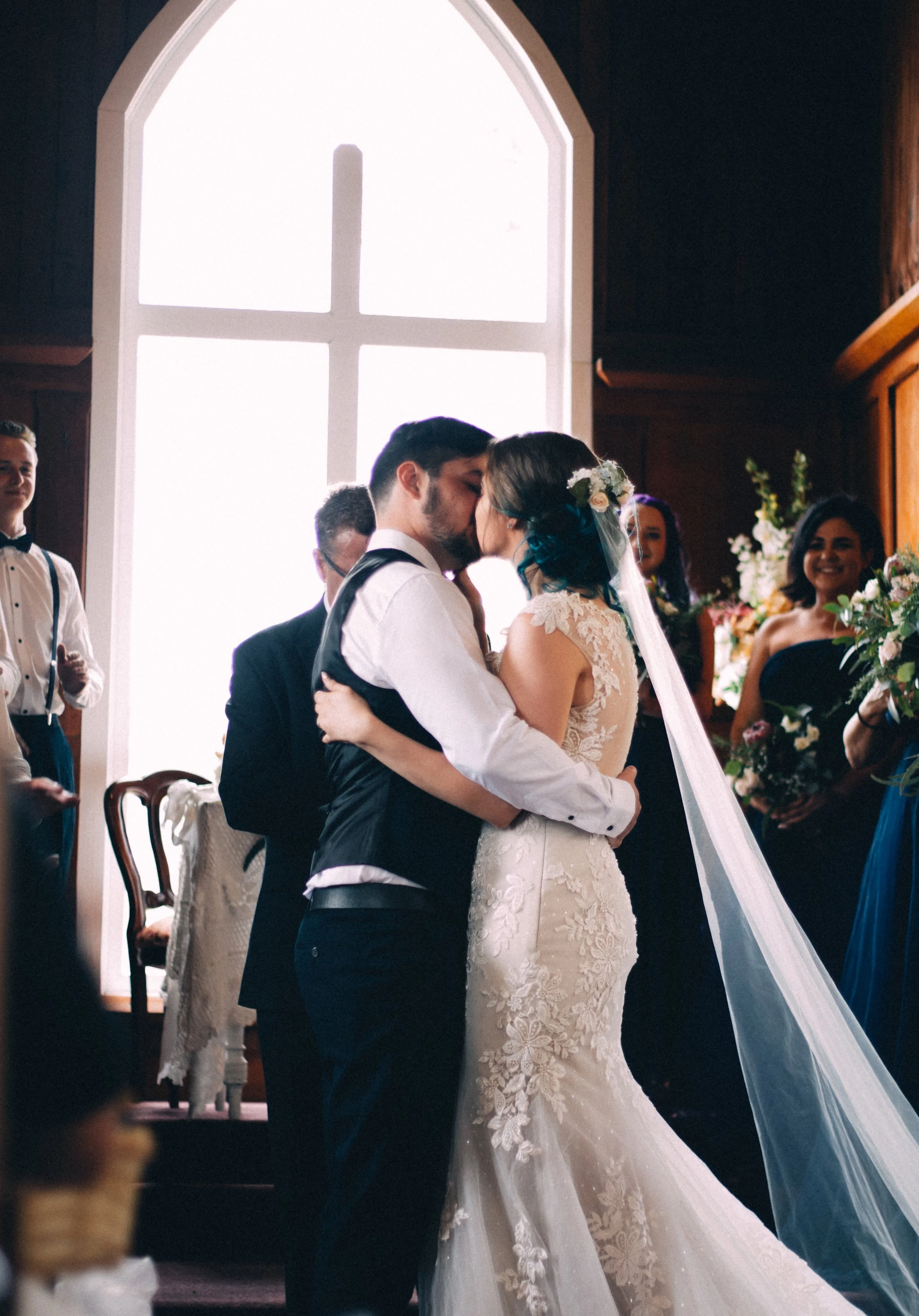 A bride and groom share a kiss during their wedding ceremony inside a church. The bride wears a lace wedding gown and veil adorned with flowers, and the groom wears a black vest and white shirt. Guests stand nearby, and large windows let in bright light.