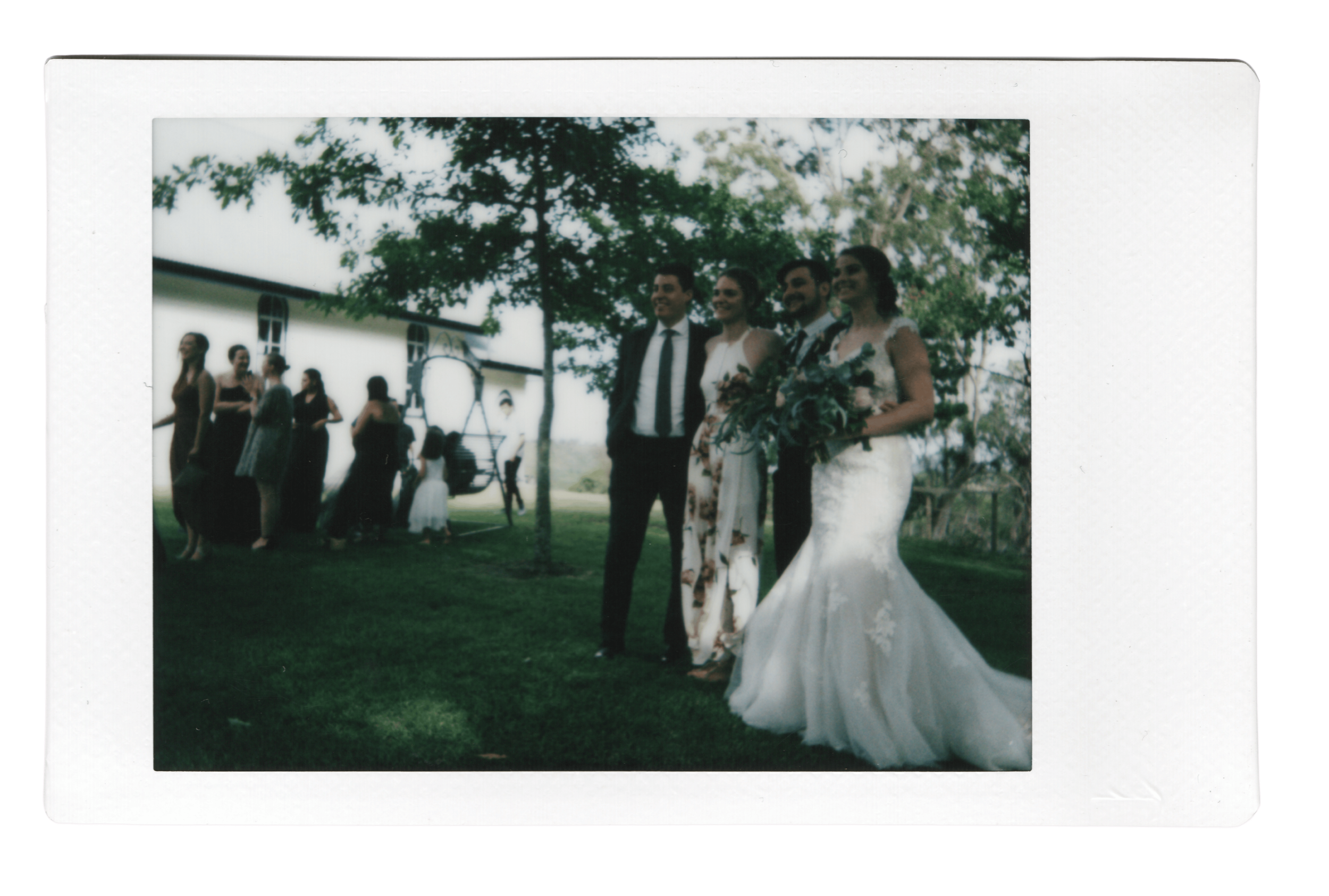 Group of people, including a bride in a white wedding gown holding a bouquet, standing outdoors under a large tree during a wedding ceremony or celebration.