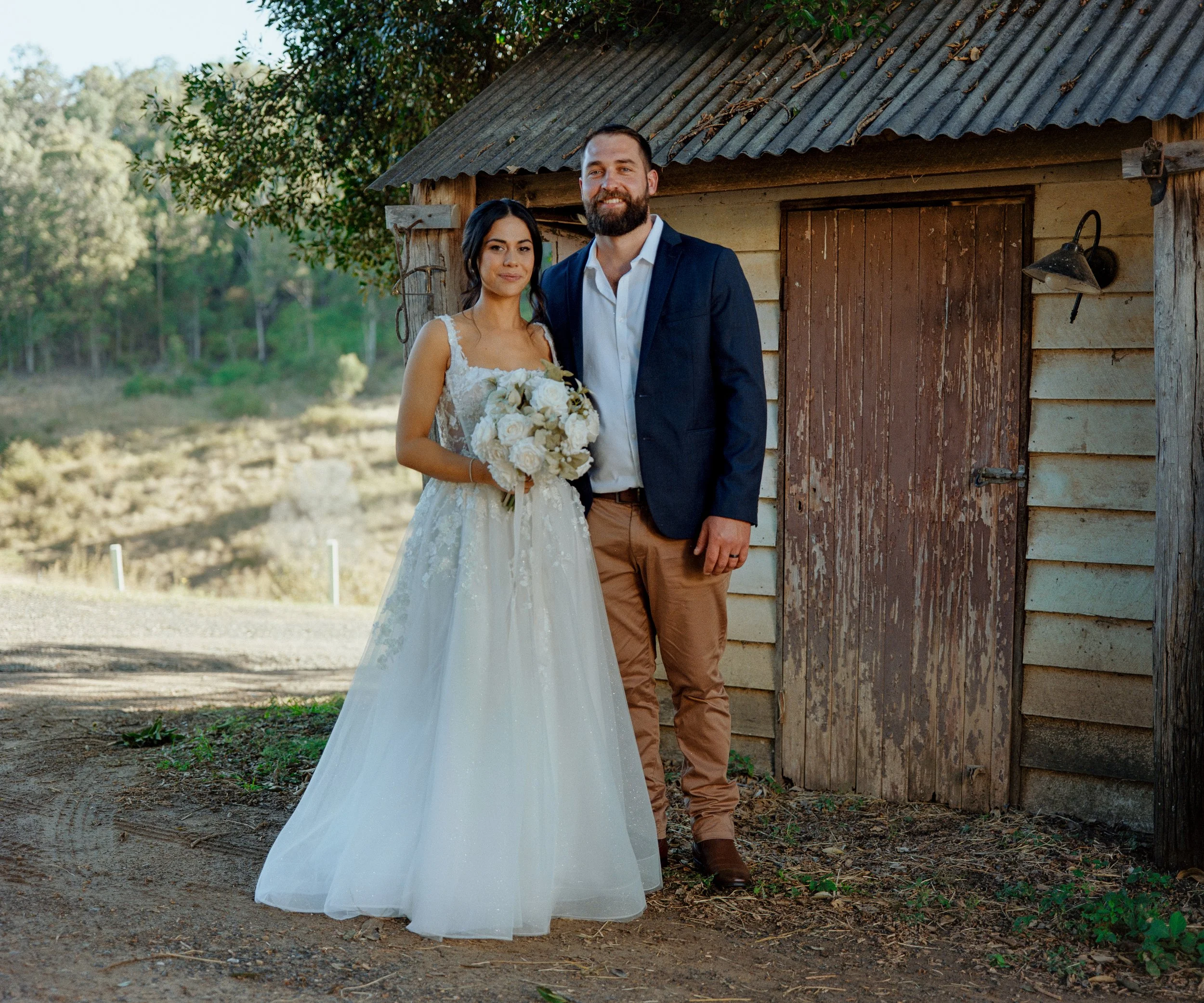A bride and groom standing outside in front of a rustic wooden shed, smiling at the camera on their wedding day. The bride is holding a bouquet of white roses, and they are dressed in wedding attire with an outdoor natural setting in the background.