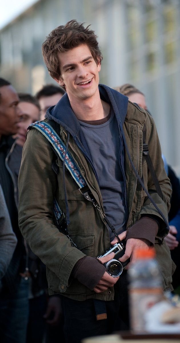 Young man with curly hair smiling, wearing a green jacket and gray hoodie, holding a vintage camera, standing outdoors among a group of people.