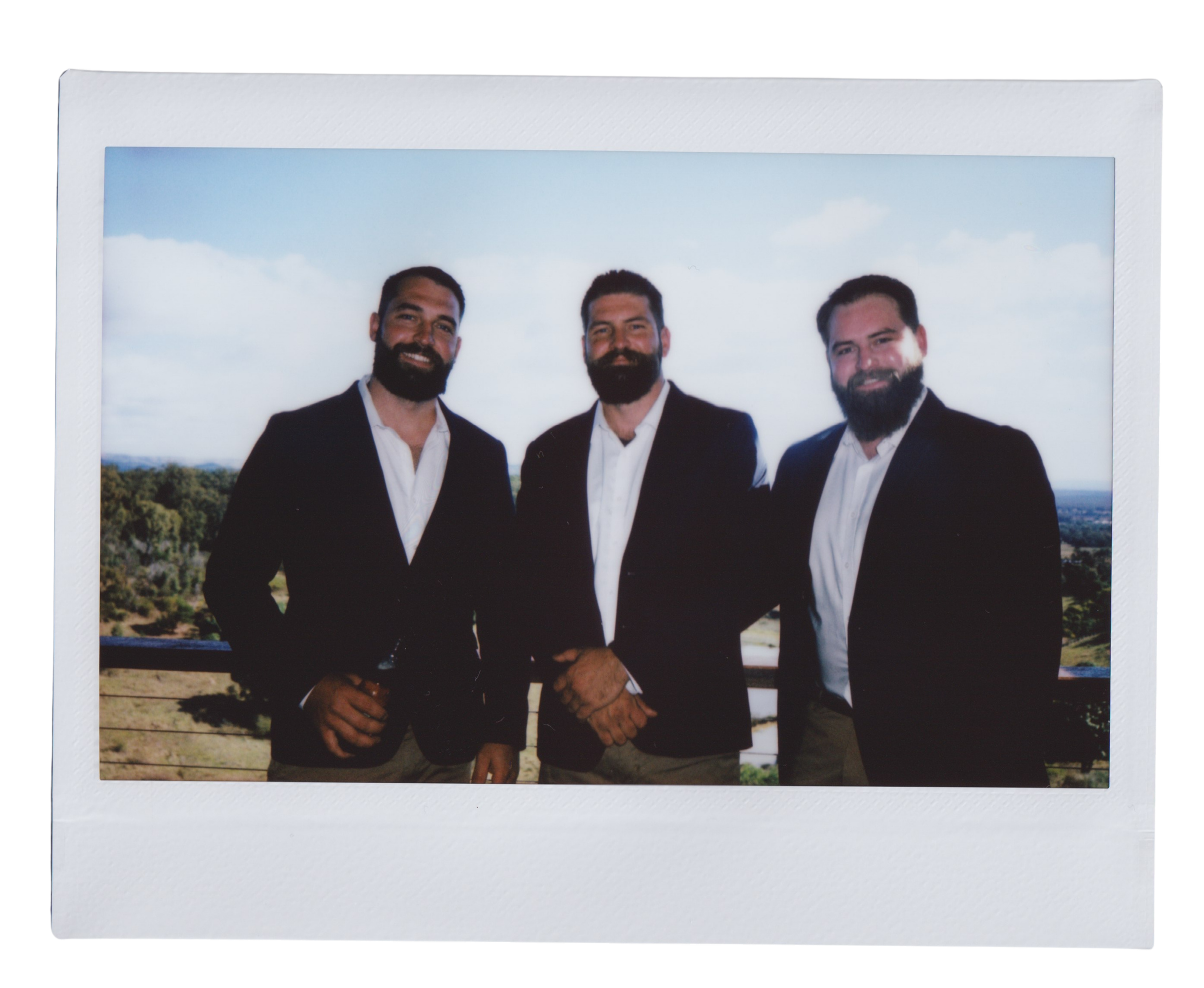 Three men in suits smiling outdoors, holding hands in front of a landscape with trees and a blue sky.