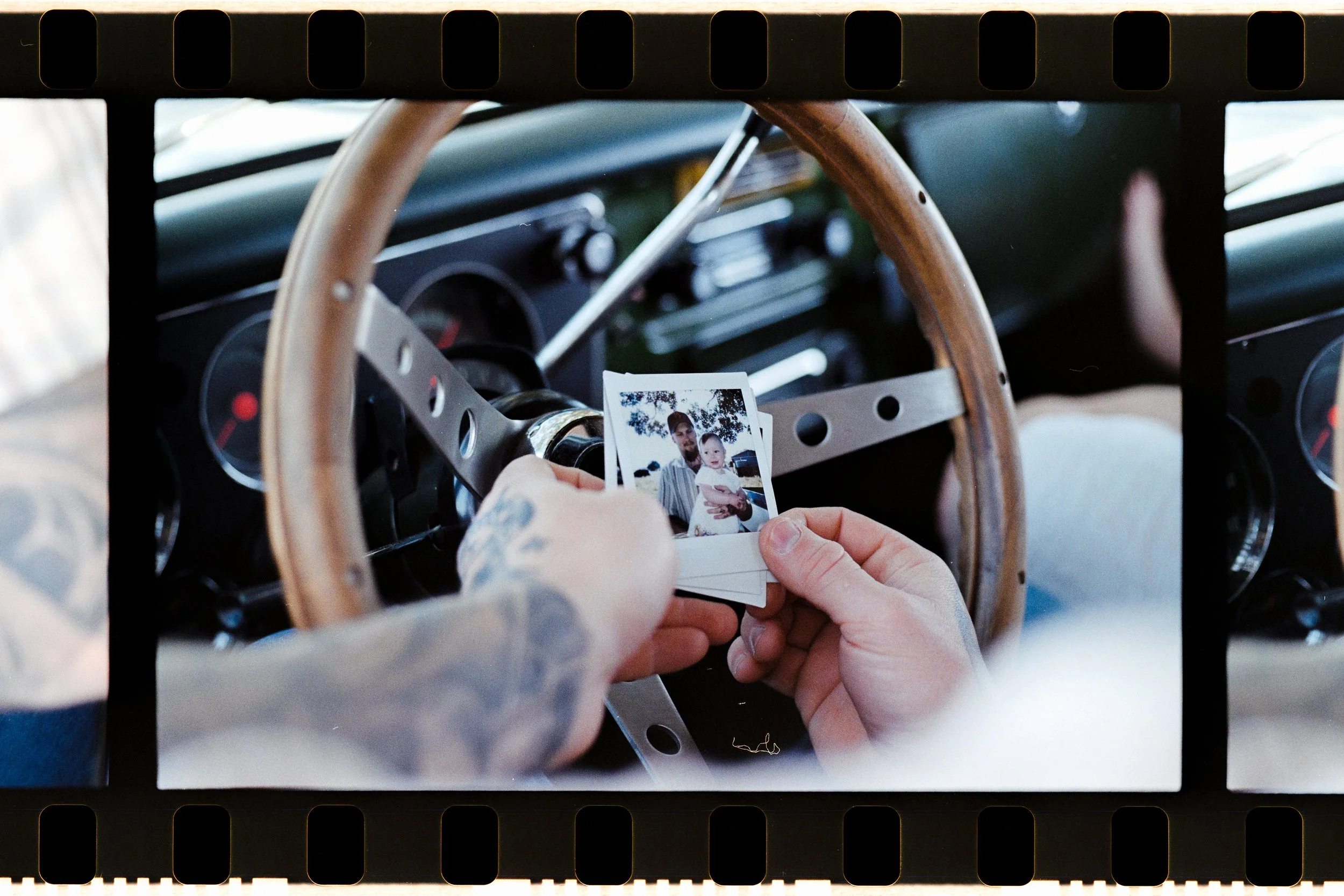 Hands holding a stack of photographs inside a vintage car with a wooden steering wheel.