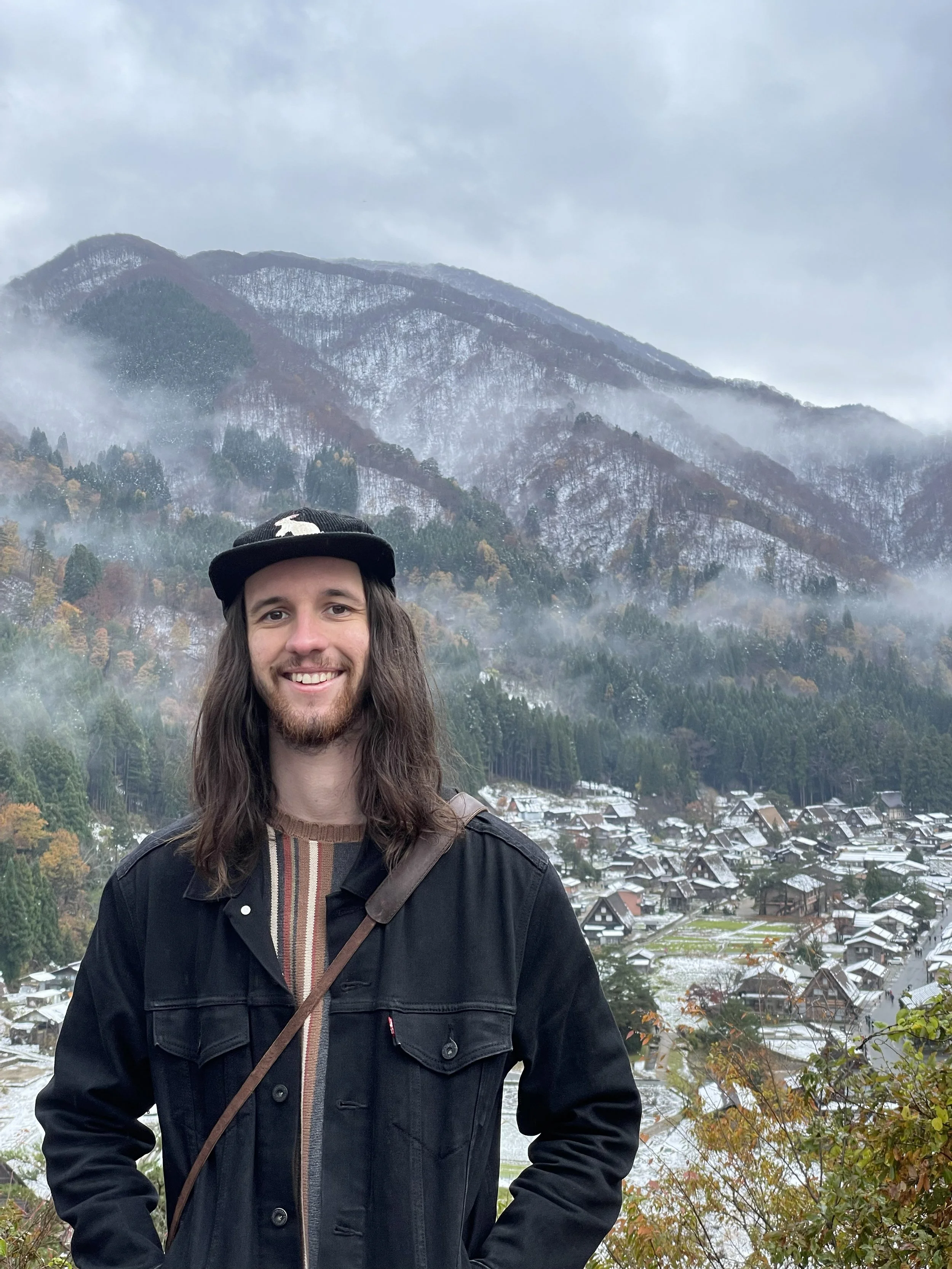 A person with long brown hair, beard, and wearing a black hat and jacket, smiling in a mountainous landscape with a village below on a cloudy day, with snow on the rooftops and mountains in the background.